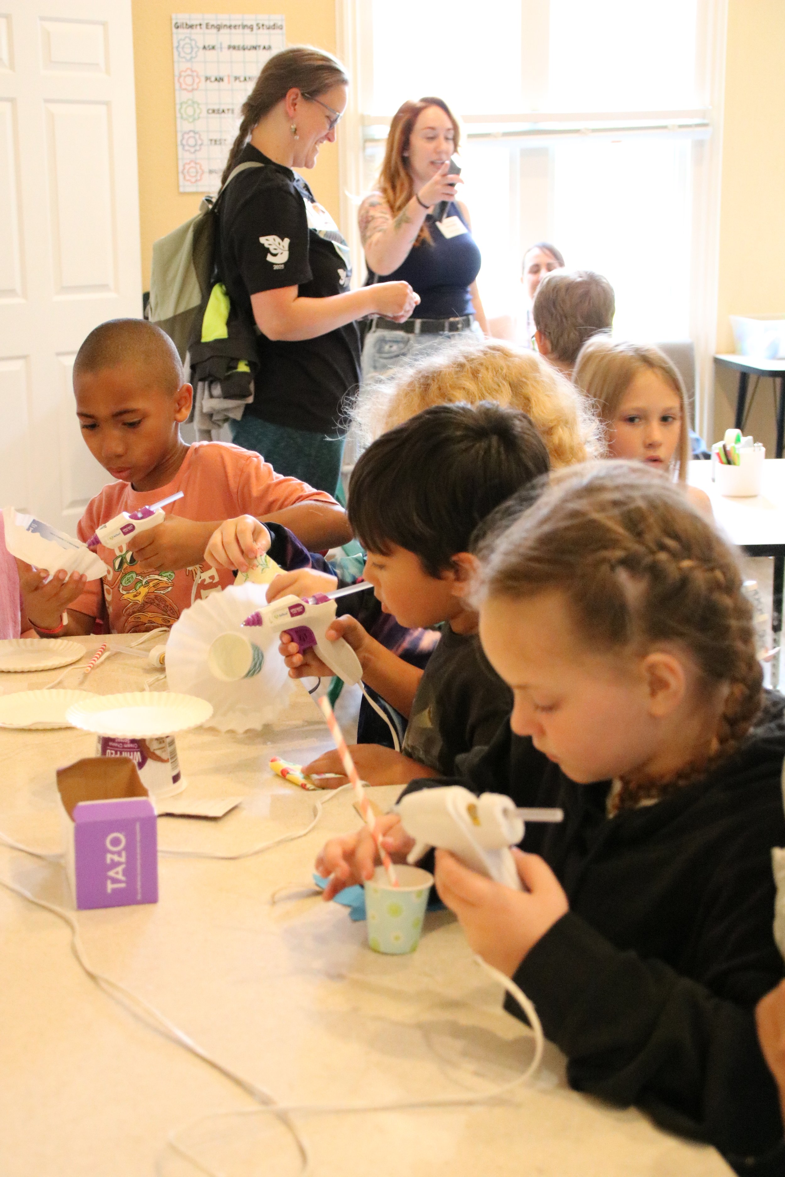 Children sitting at a table, using glue guns to decorate cupcakes, with adults standing in the background, in a classroom or activity room.
