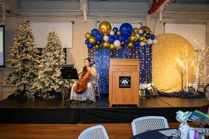 A woman playing a cello on a decorated Christmas-themed stage with two snow-covered Christmas trees on the left, colorful balloons, a glittery gold sphere, and white lights on the right.