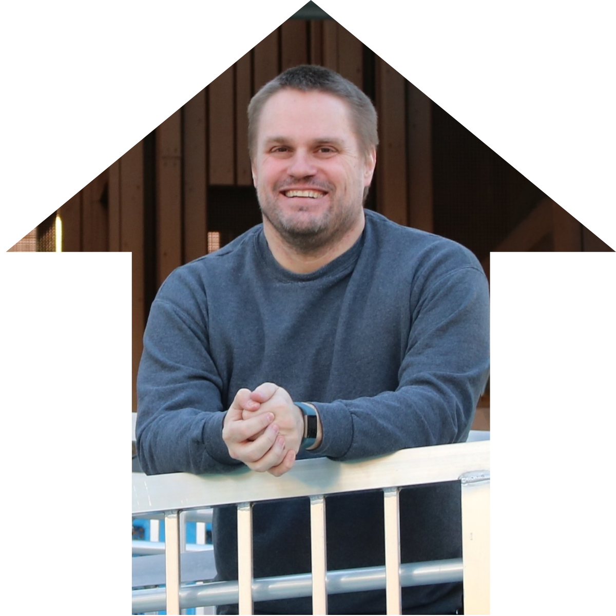 A man with short brown hair and a beard, smiling and sitting at a table outdoors before a wooden fence.