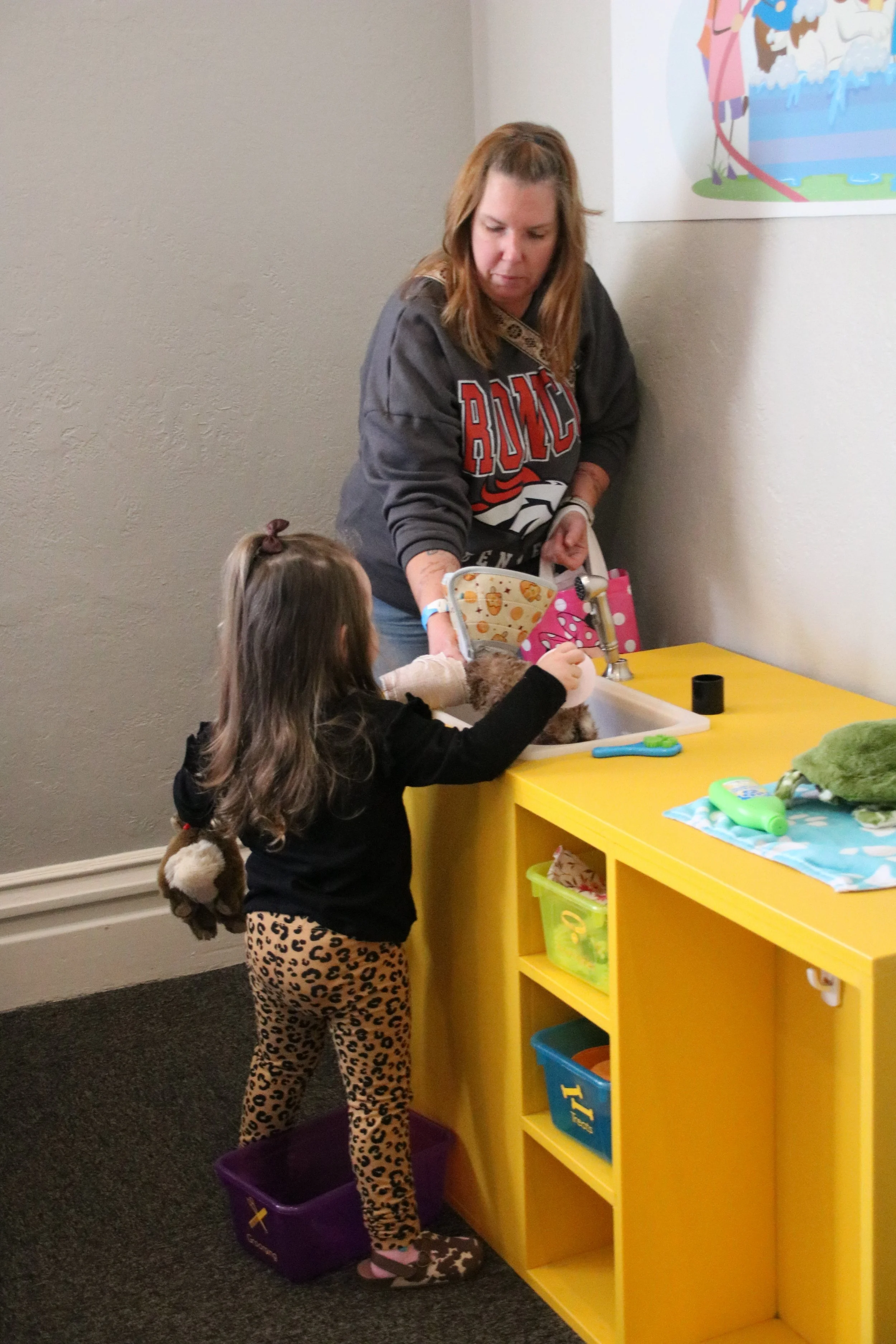 A woman and a young girl playing with a small dog in a pretend veterinary clinic, with toys and sanitized supplies on a yellow counter.