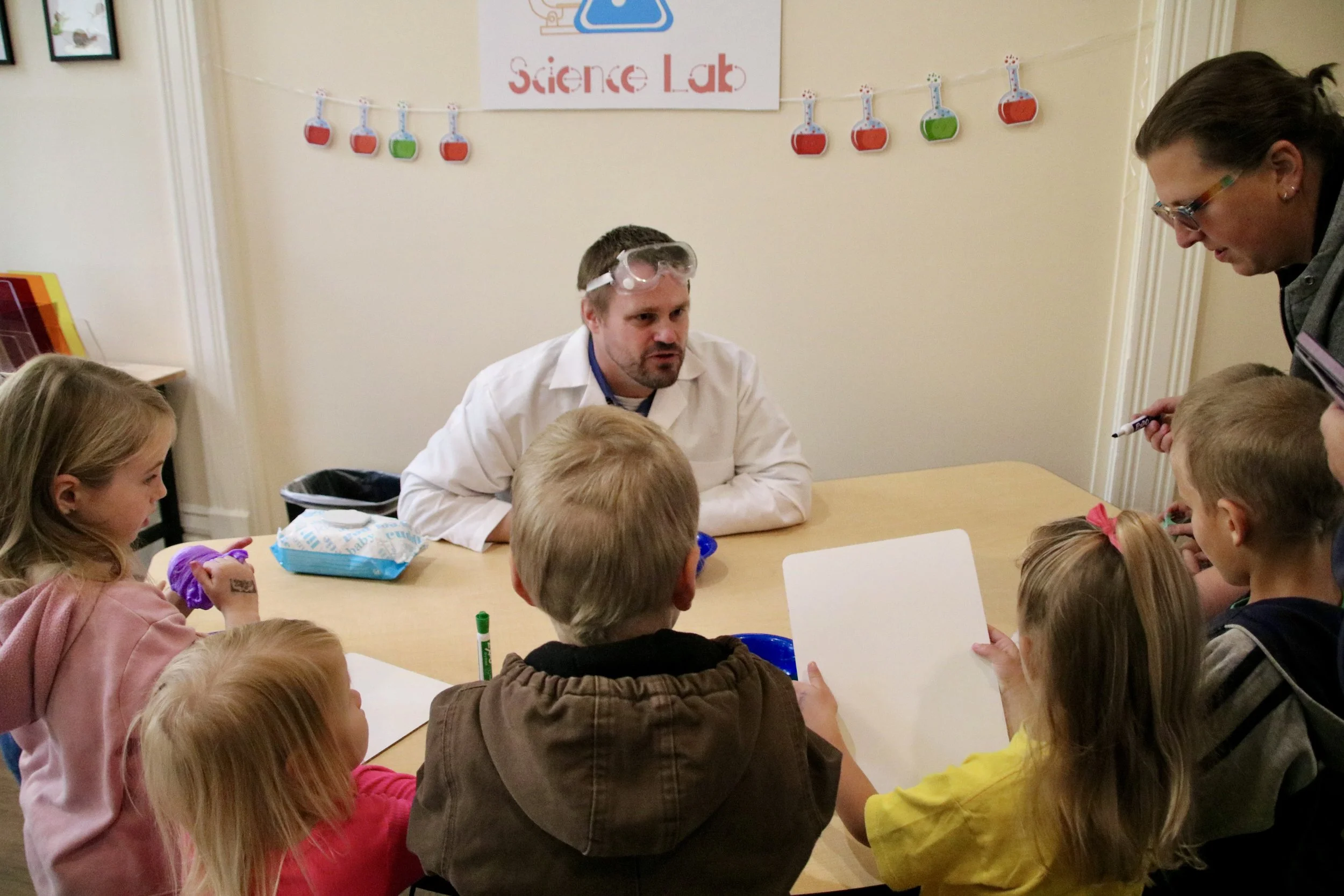 A science teacher in a lab coat and goggles talking to five children at a science lab table, with a poster reading 'Science Lab' behind them.