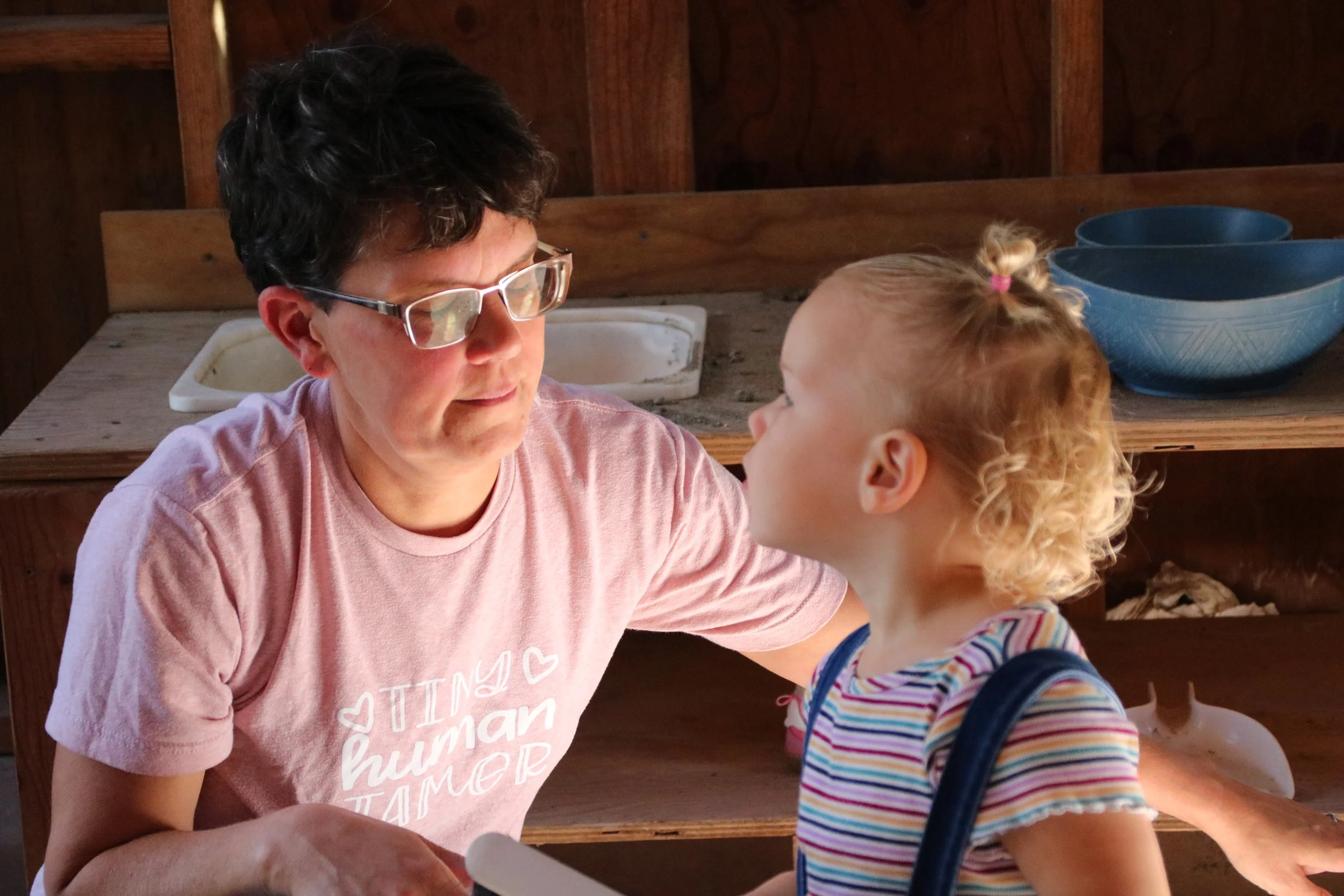 A woman with short dark hair, glasses, and a pink T-shirt with the words 'human' and 'dinner' written on it, faces a young girl with curly blonde hair in a striped shirt. They appear to be in a rustic wooden setting, possibly a kitchen or craft area, engaged in a close interaction.