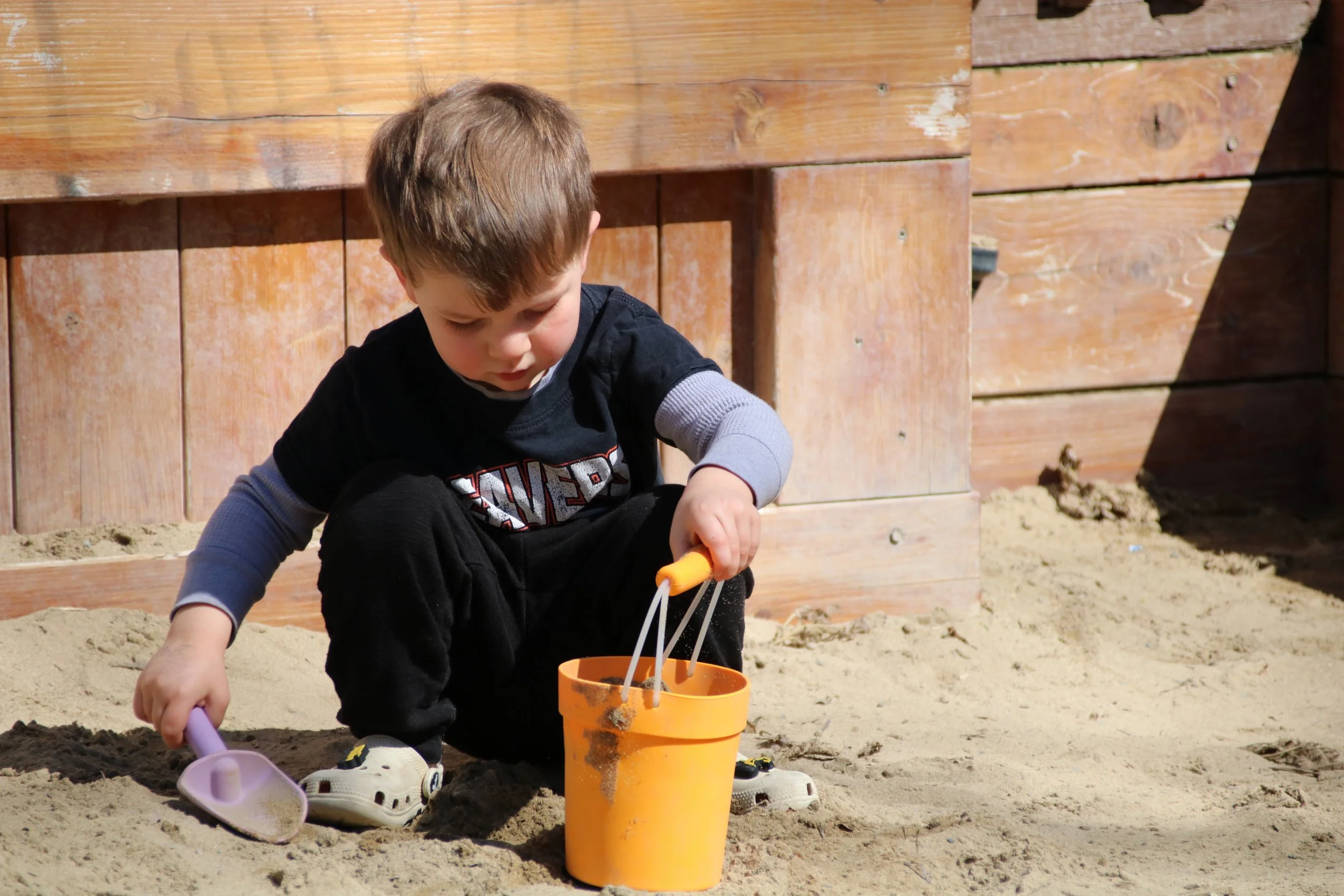 A young boy playing in a sandbox with plastic shovels and a bucket, with a wooden fence in the background.