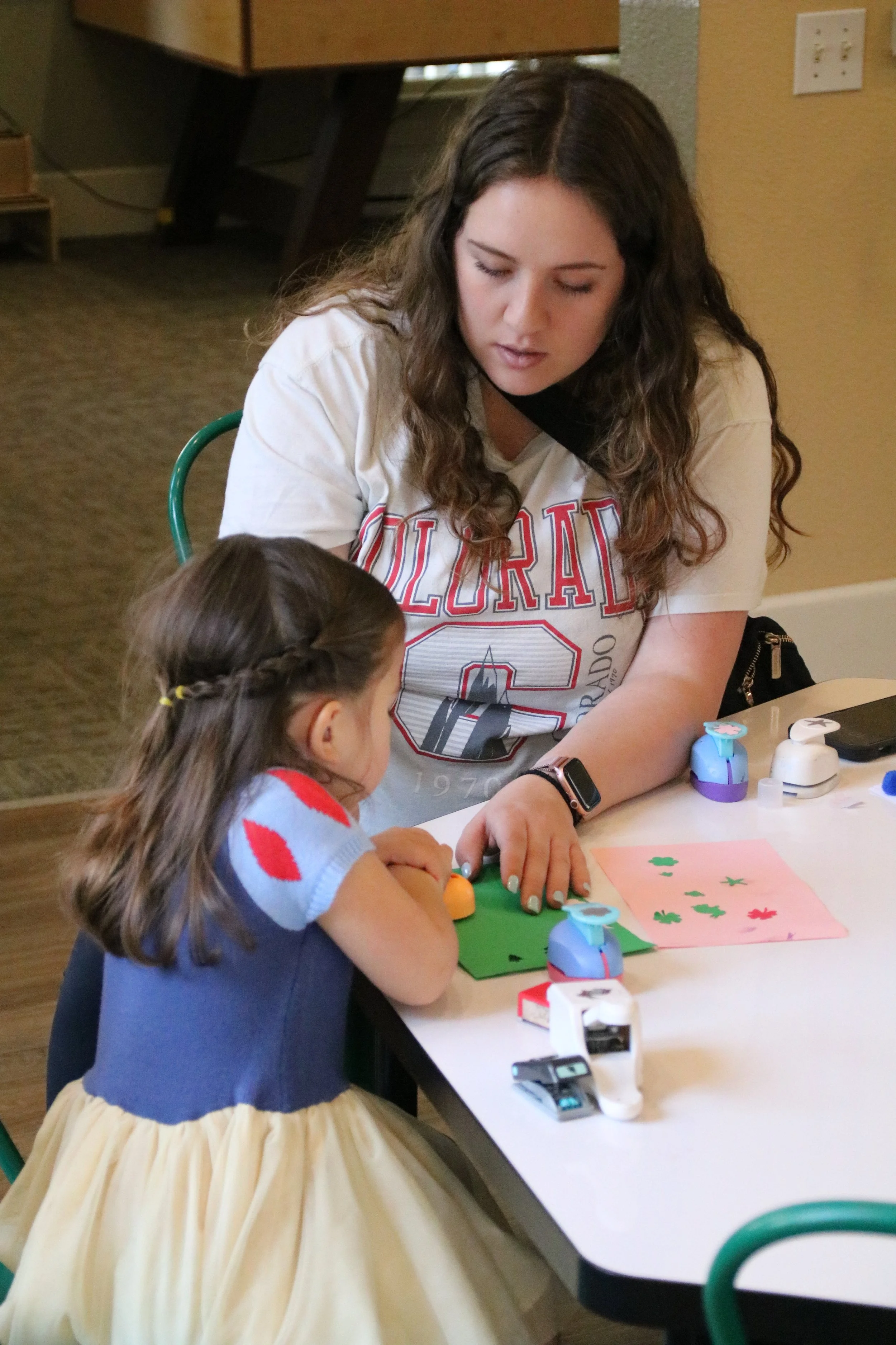 A woman showing a young girl how to make a craft project with colored paper, stamps, and stickers at a table.