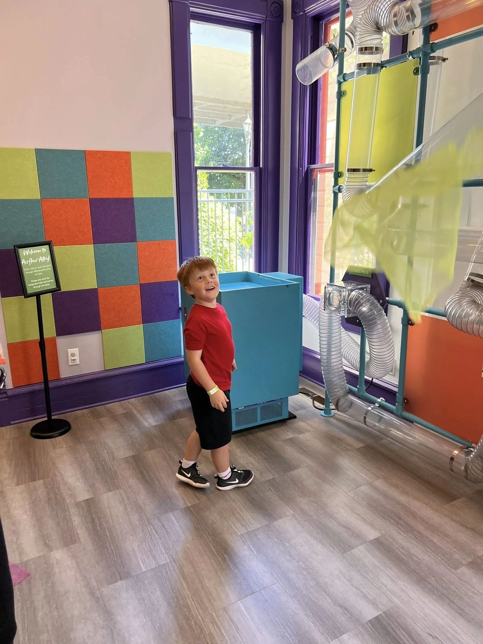 A young boy in a red shirt and black shorts standing in a colorful indoor space with purple, orange, green, and blue walls, large windows, and a mechanical structure with clear tubes and vents.