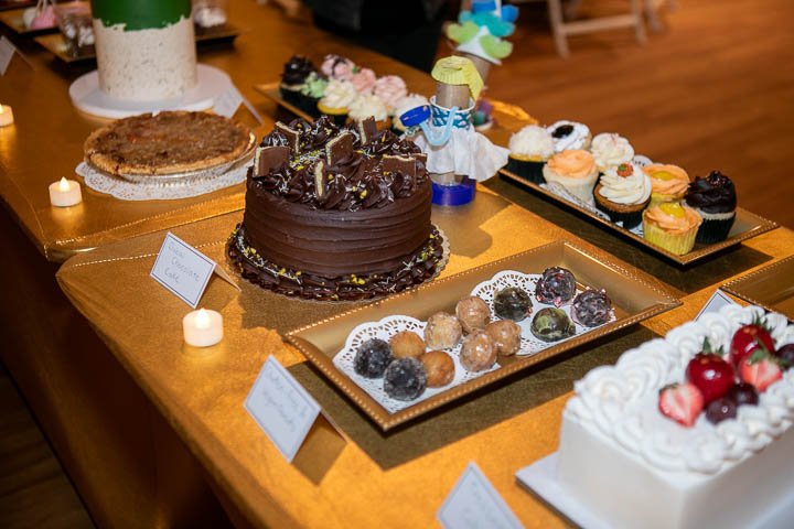 A table with a variety of desserts including a large chocolate cake, cupcakes, fruit-topped cake, and assorted cookies and chocolates, decorated with small candles and a decorative figure.