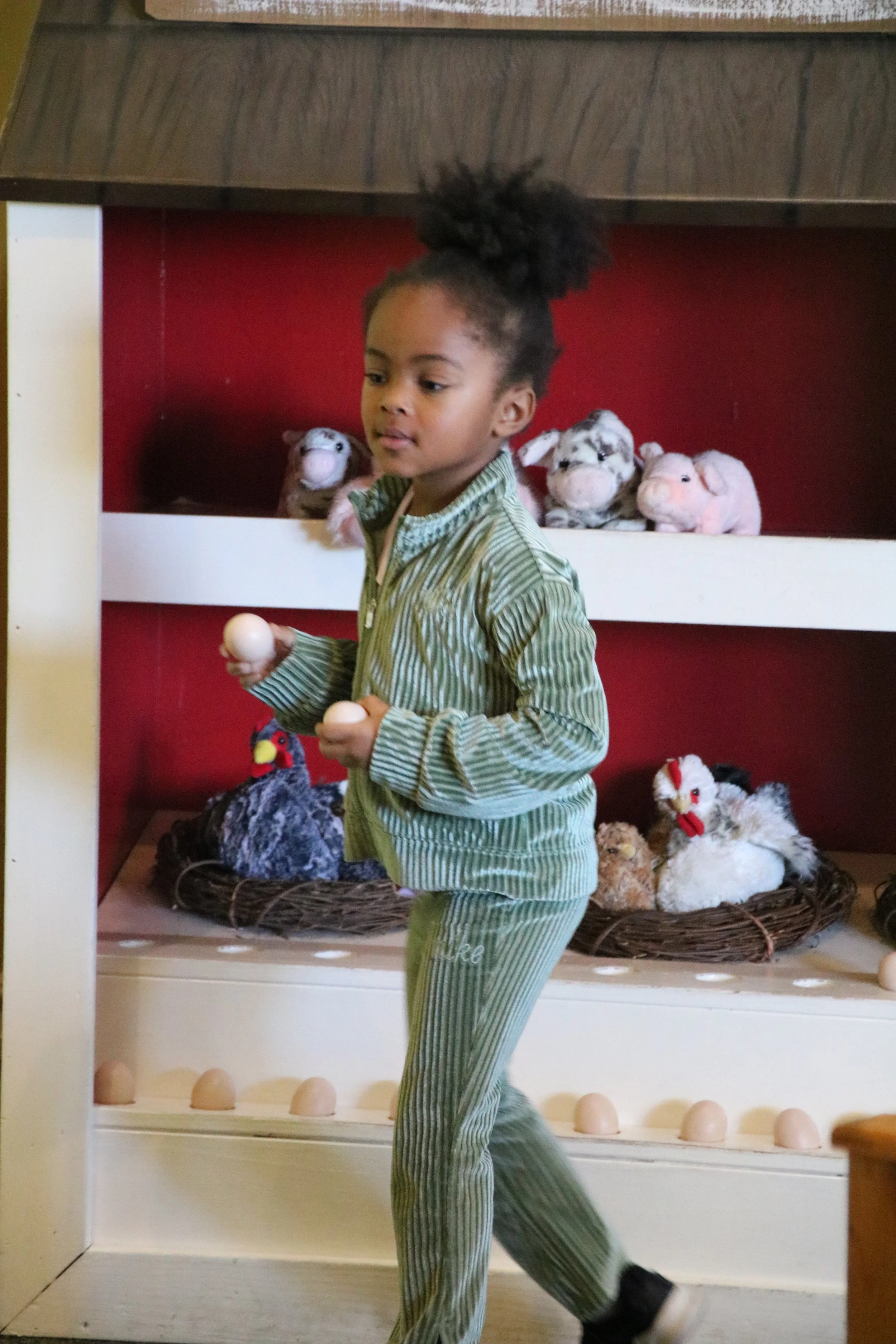 A young girl in green striped pajamas holding a small white ball stands in front of a shelf with stuffed animals, including plush dolls and chickens.