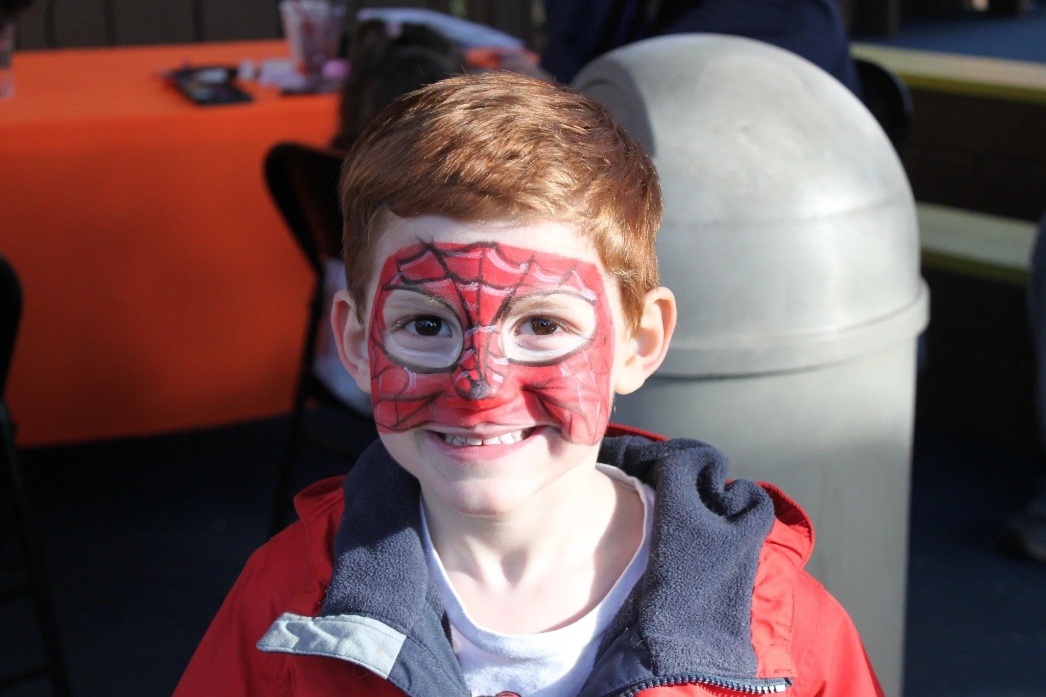 A young boy with face paint resembling Spider-Man's mask, smiling at the camera, outdoors.