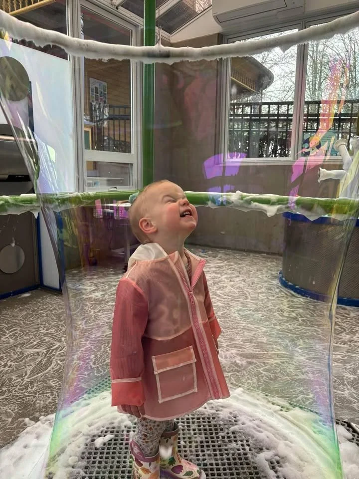 A young girl with a blonde bun standing inside a large, colorful soap bubble, crying or upset while looking upward. She is wearing a pink and beige jacket, patterned leggings, and rain boots. The background shows a room with windows, pipes, and outdoor scenery.