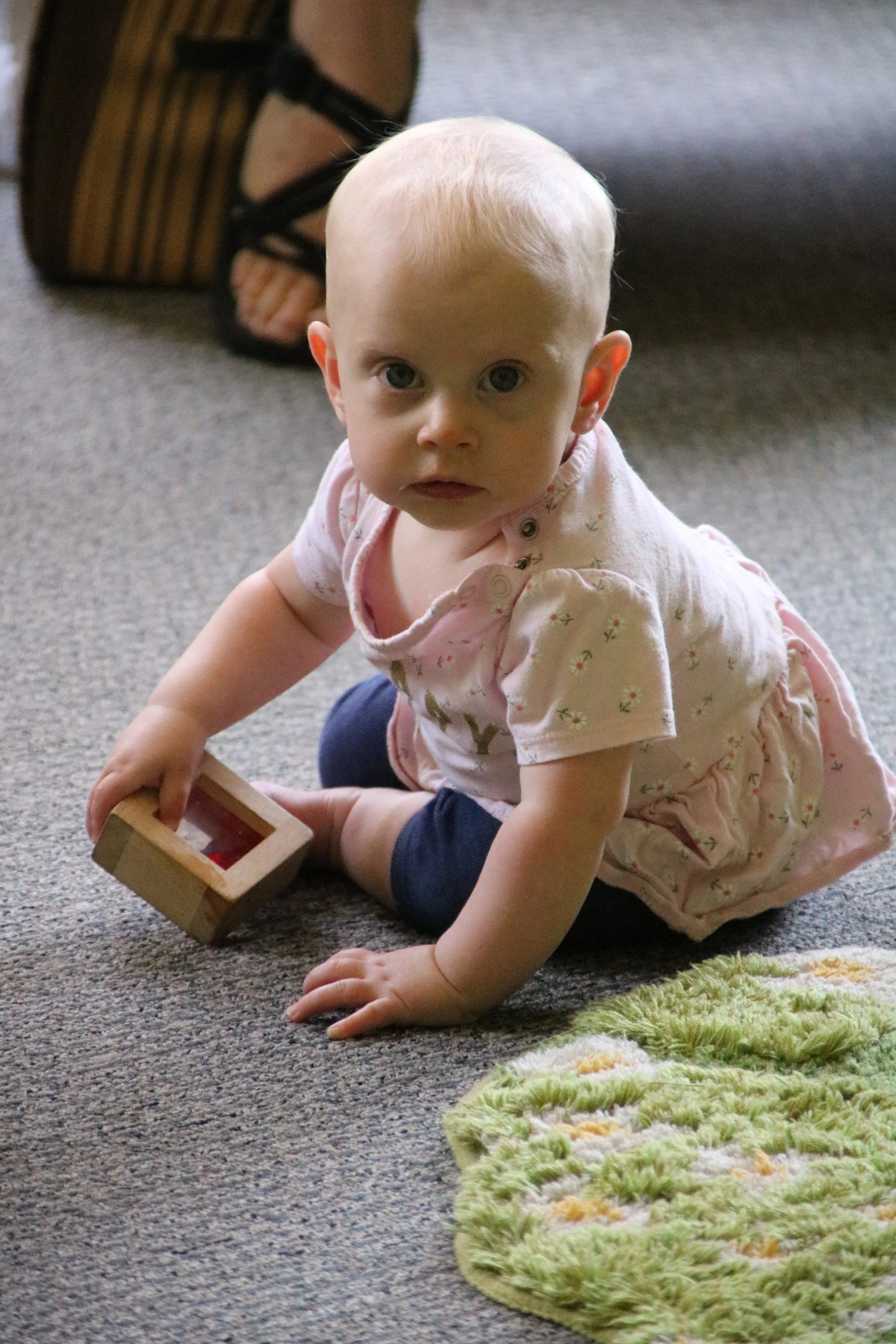 A young toddler girl with blonde hair, sitting on a carpeted floor, holding a wooden toy, and looking up at the camera with a curious expression.