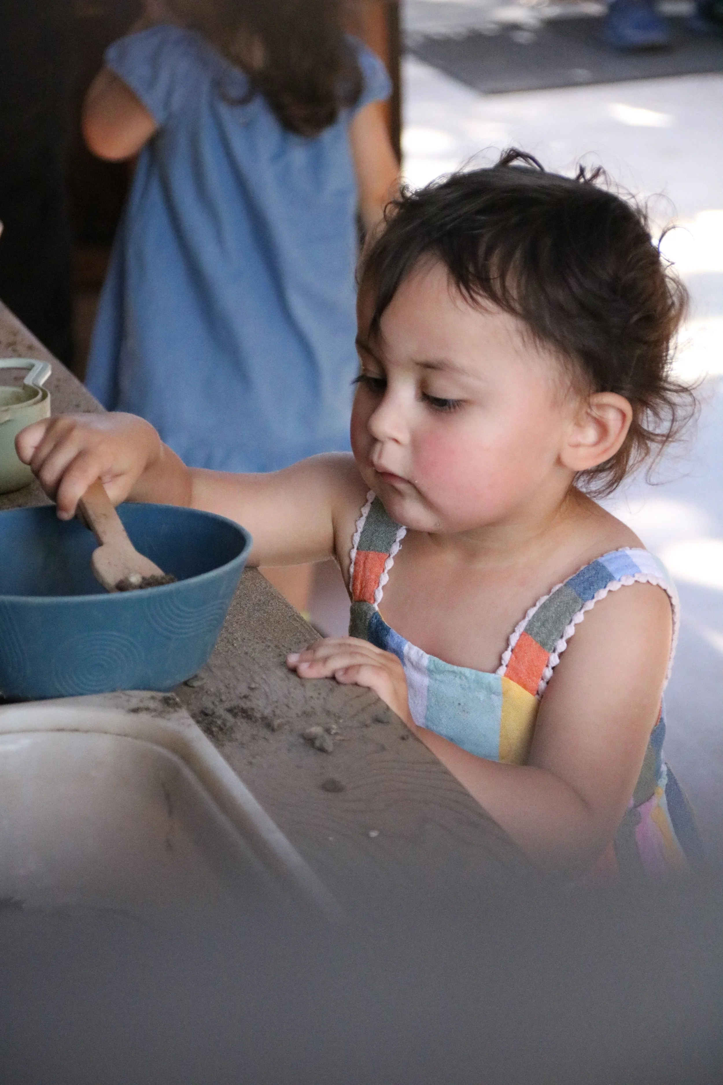 Young girl with dark hair, wearing a colorful plaid dress, playing with dirt in a blue bowl at an outdoor setting.