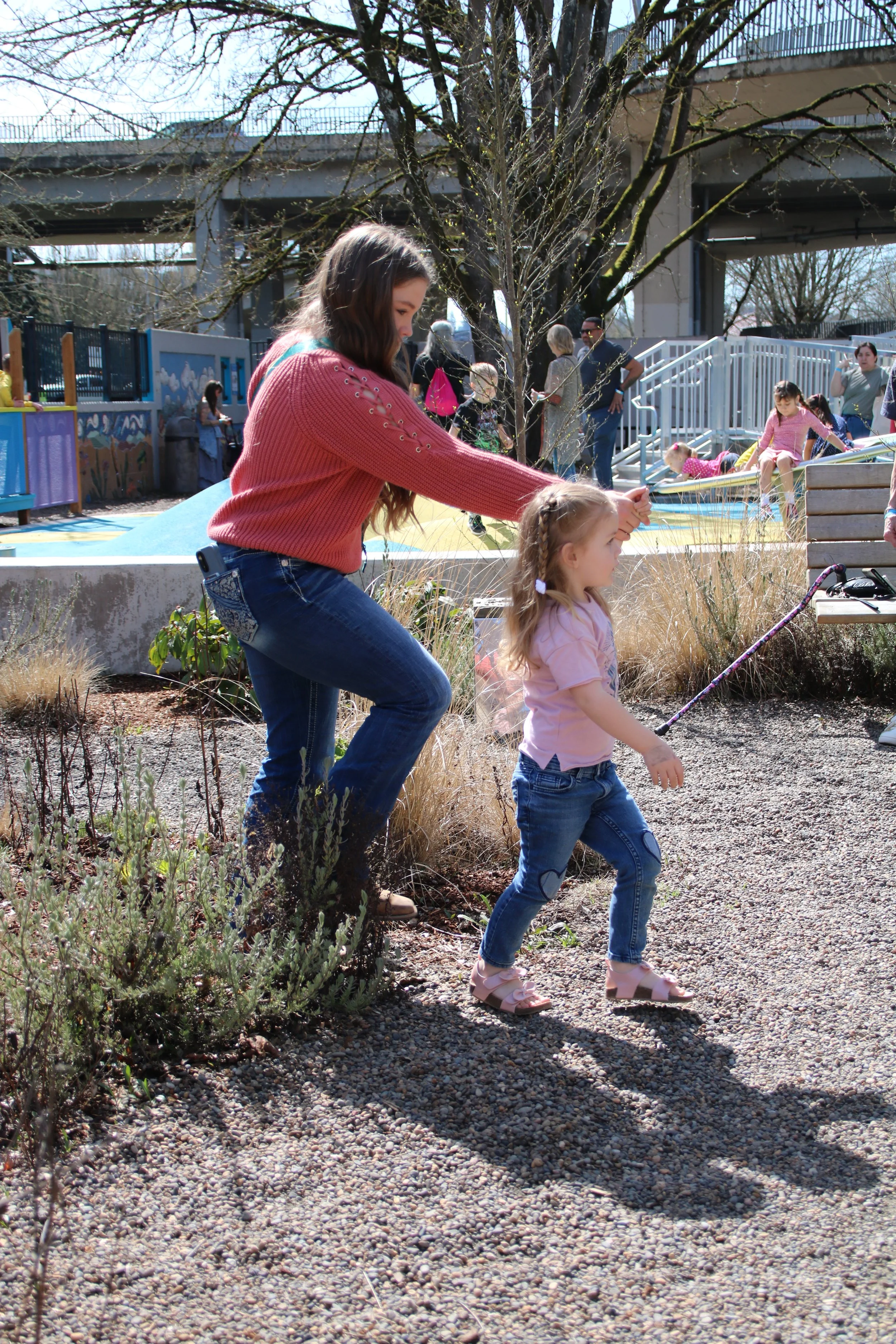 A young woman helping a little girl walk on a gravel path in a park, with a playground and other people in the background during daytime.