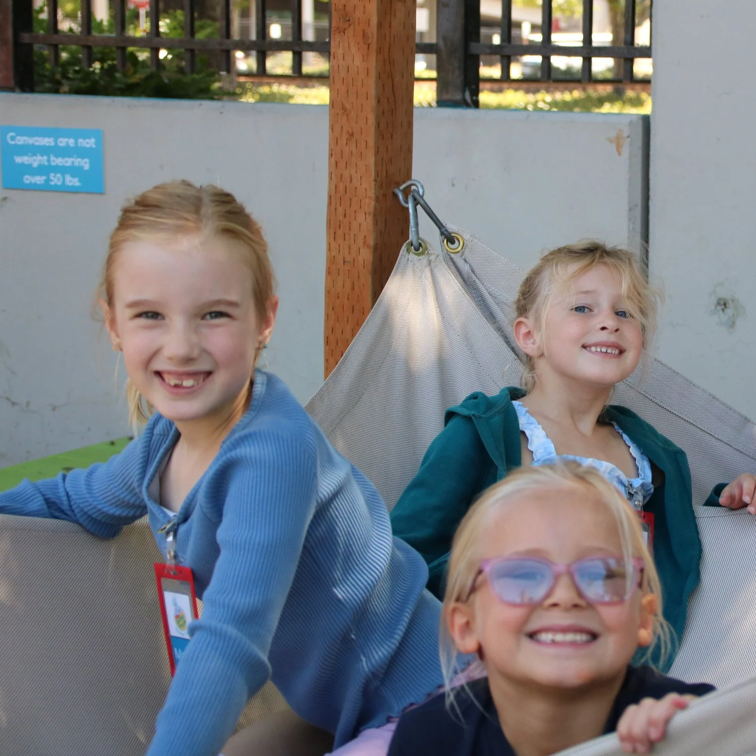 Three young girls smiling and playing on a playground structure with a metal and fabric design, outdoors with a concrete wall and a blue informational sign in the background.