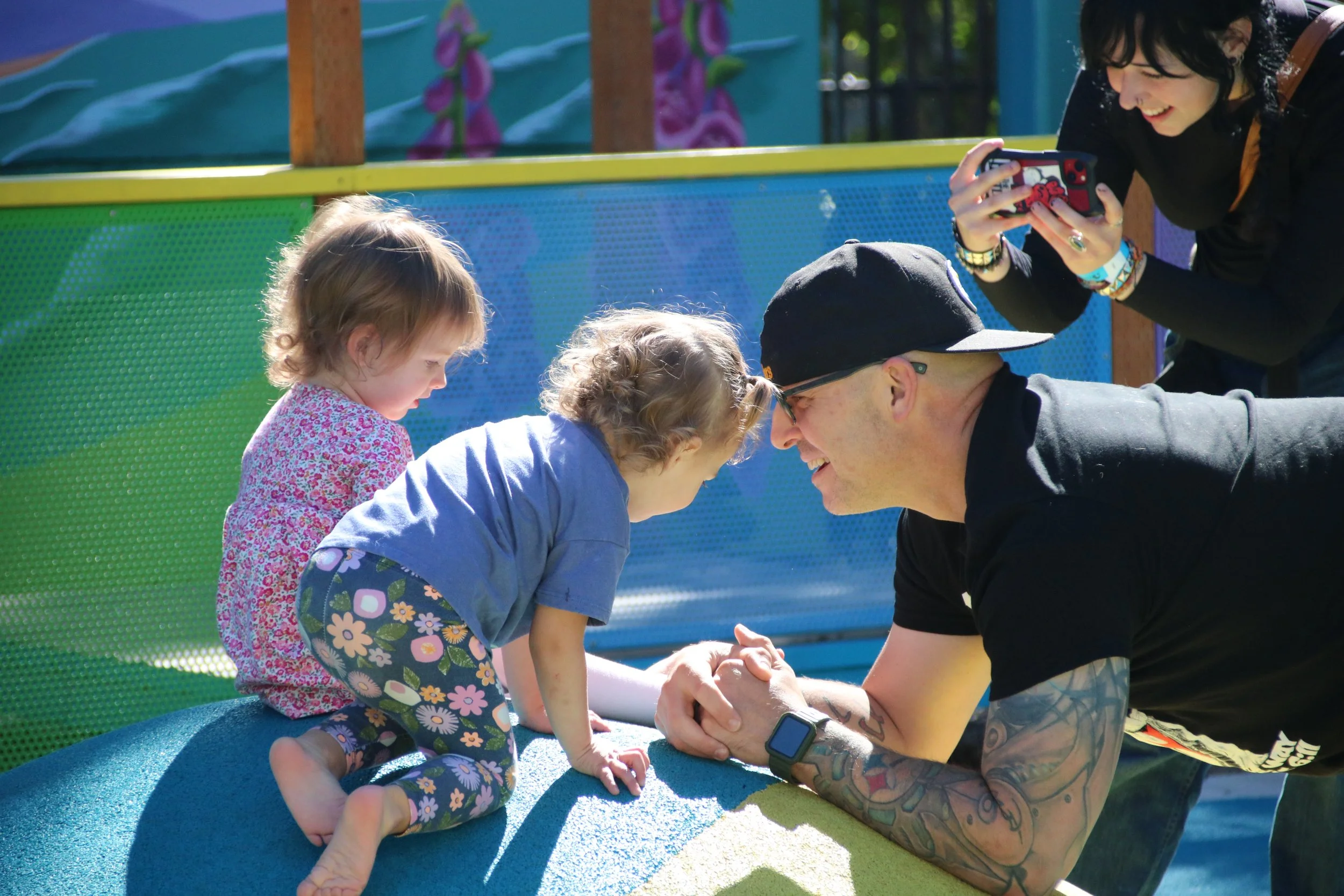 Two young girls are playing on a colorful playground with an adult man, who is leaning on the playground equipment and holding their hands. An adult woman is taking a photo of the scene with her smartphone.