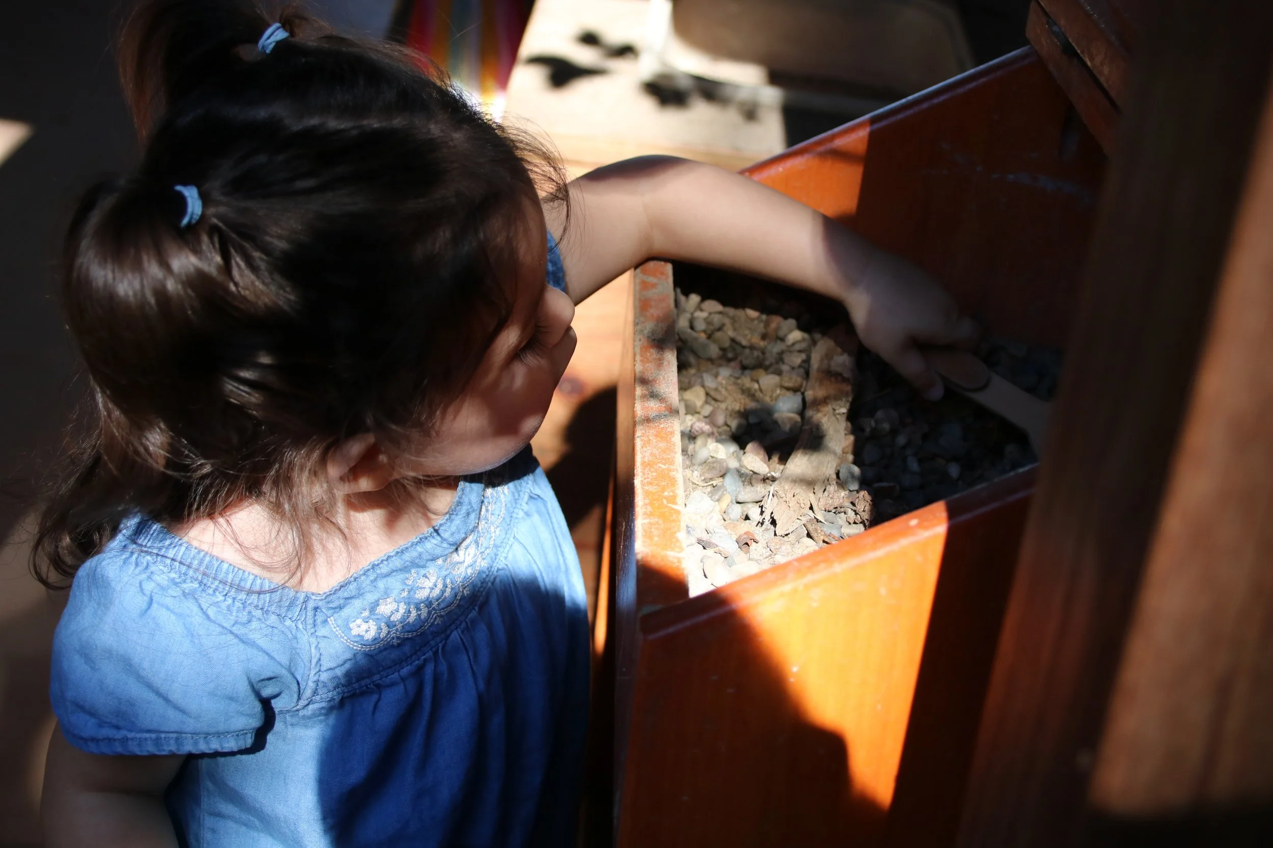 A young girl in a blue dress is reaching into a wooden box filled with small rocks and dirt, exploring its contents.