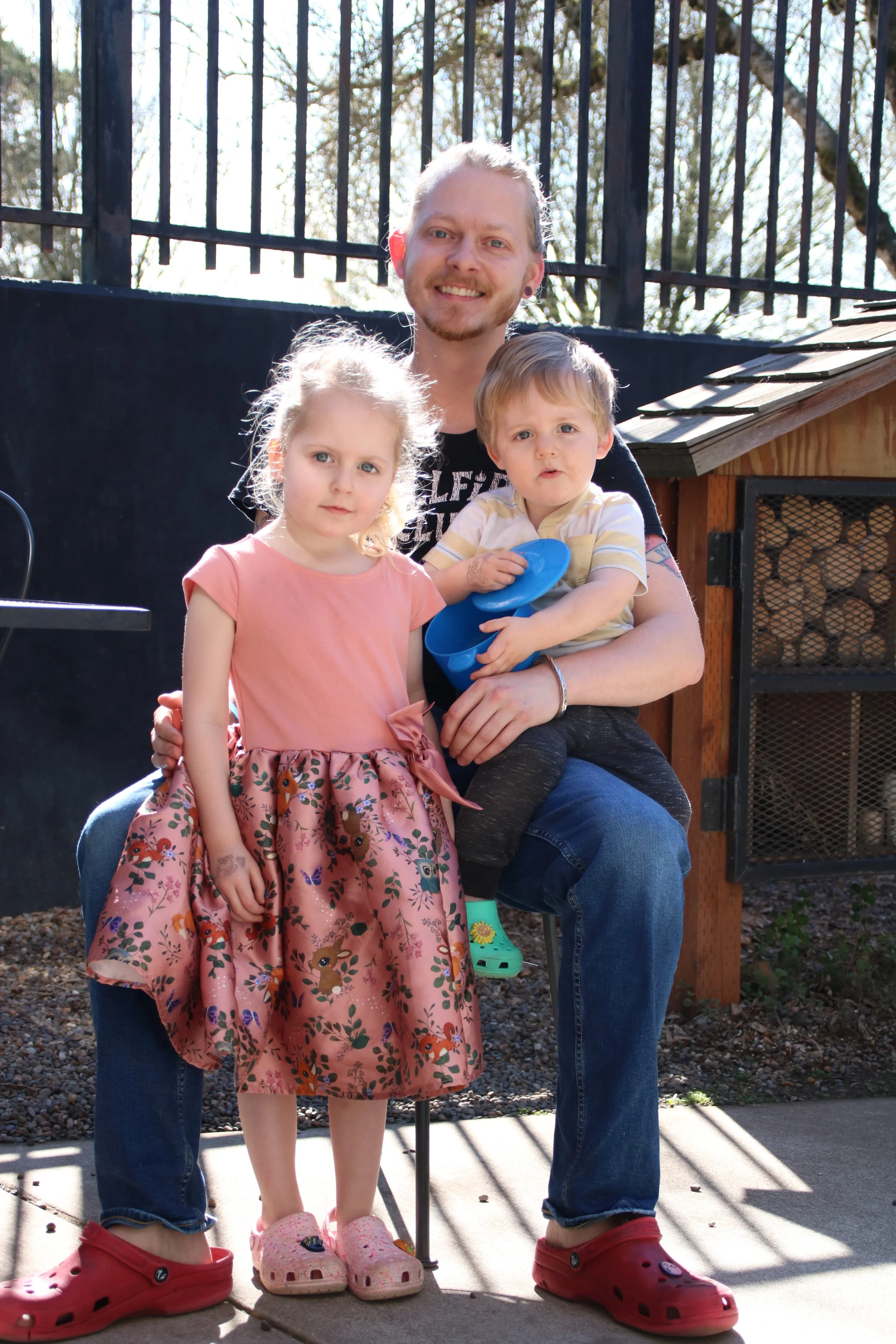 Portrait of a smiling father sitting outdoors, holding a toddler boy in a striped shirt and blue pot, next to a young girl in a pink floral dress, with a small wooden house structure behind them.