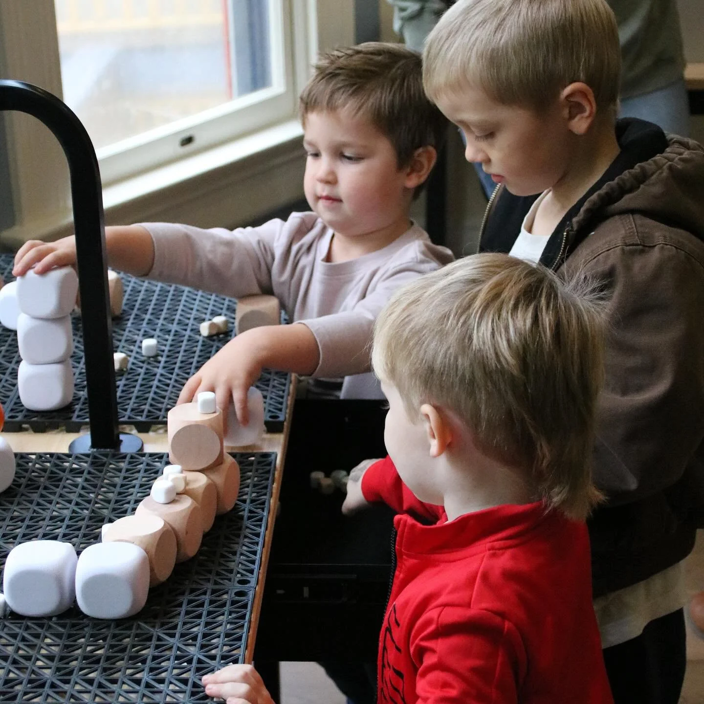 Four young boys with light skin, two with blond hair and two with brown hair, are engaged in an activity with stacking white and light pink blocks at a table near a window in a classroom or playroom.