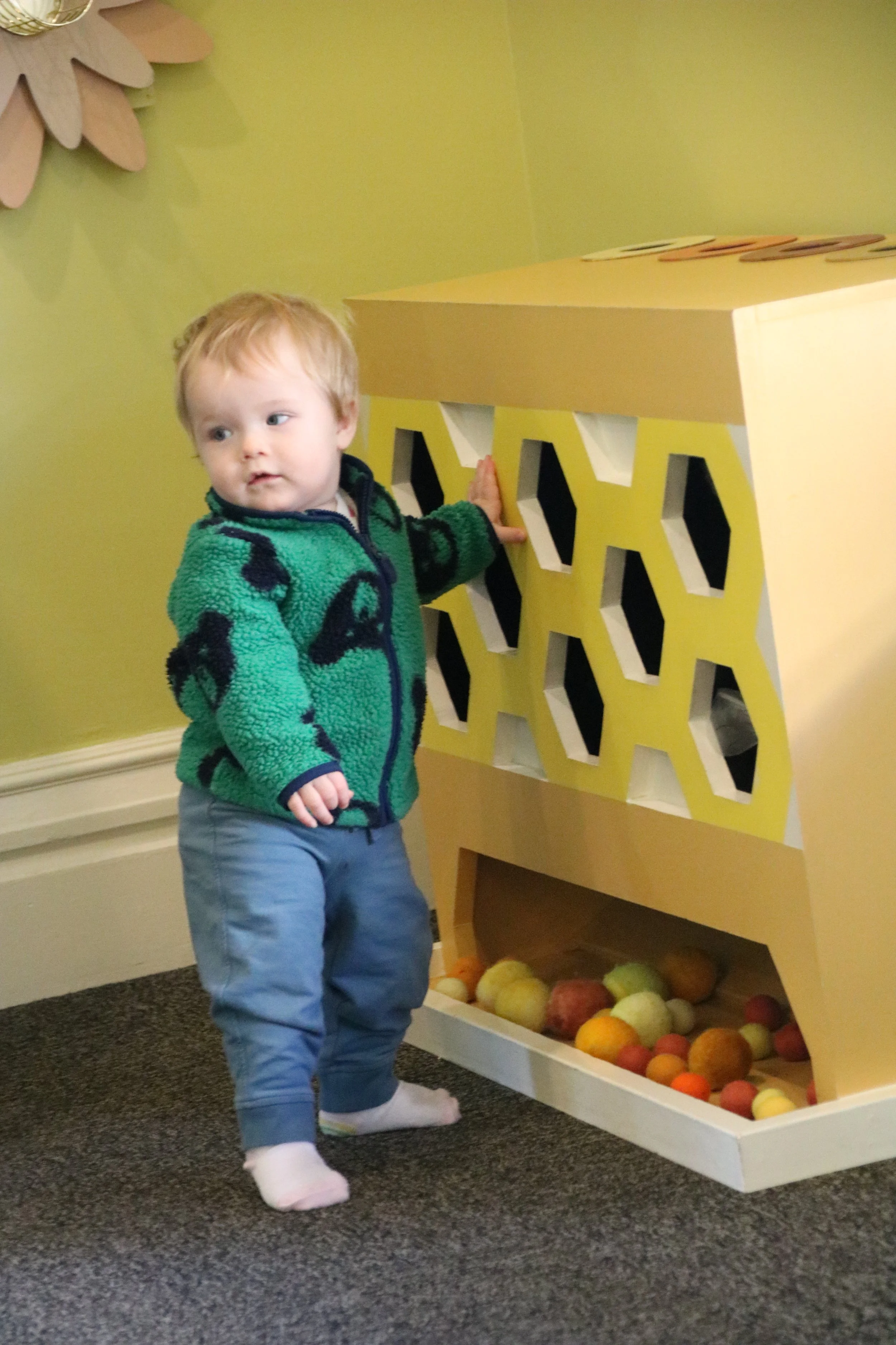 A young child with light hair, wearing a green and black jacket, gray pants, and white socks, standing next to a yellow game or display case filled with colorful felt balls, in a room with light green walls and dark gray carpet.