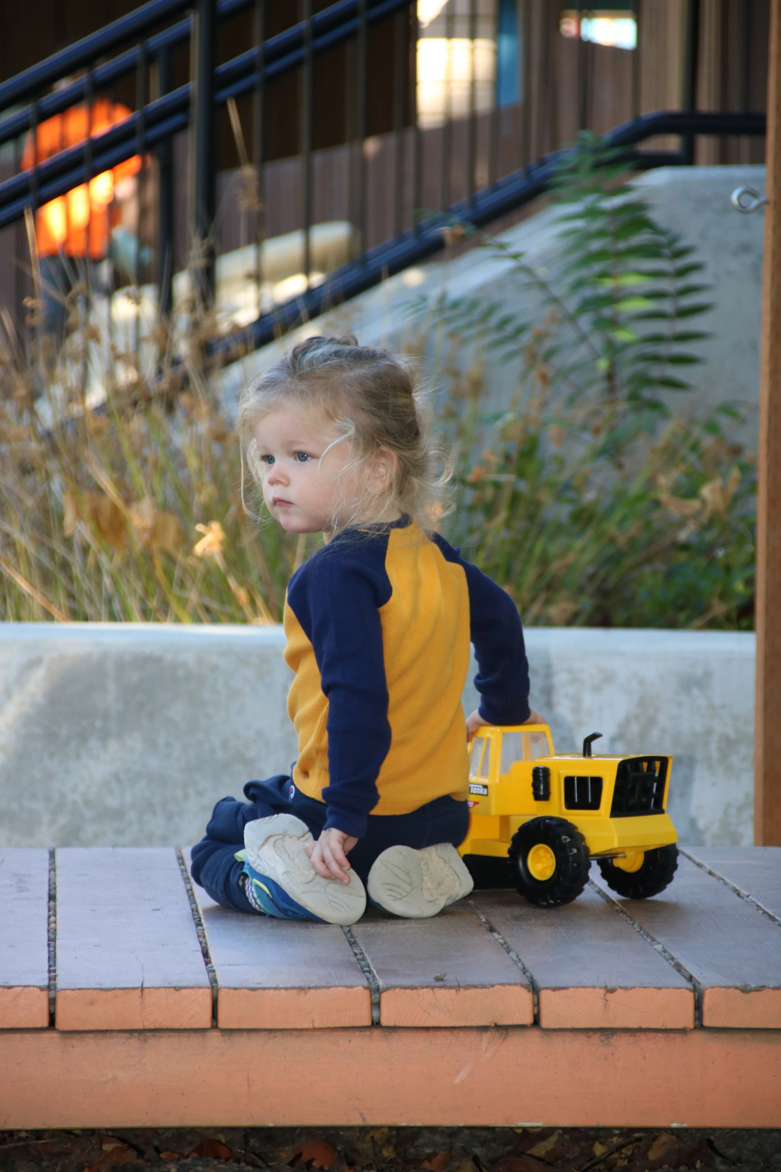 A young girl with blonde hair sitting on a wooden surface outdoors, near a yellow toy truck, with plants and a railing in the background.