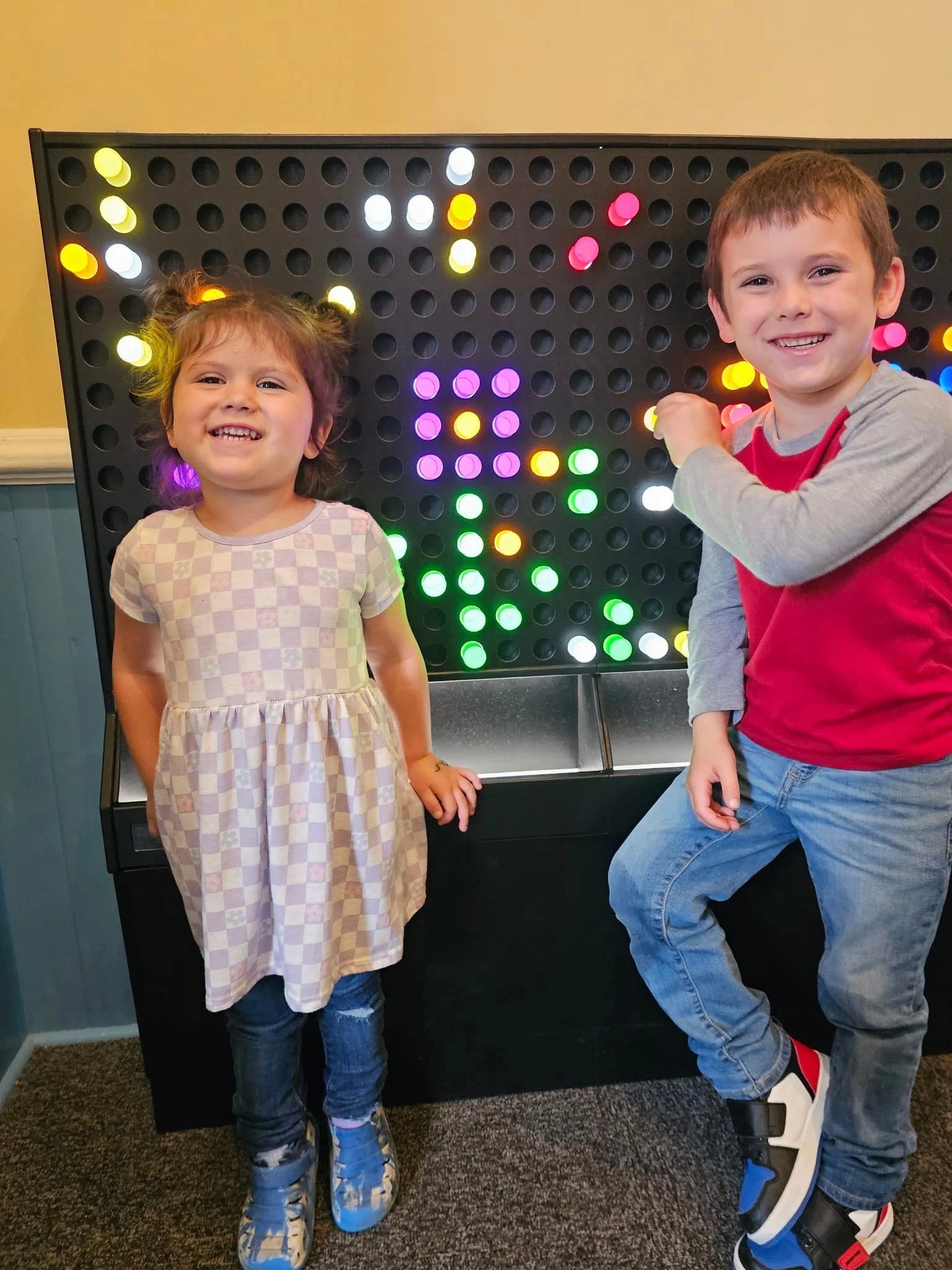 Two children, a girl and a boy, standing next to a large black pegboard with colorful lights, smiling at the camera.