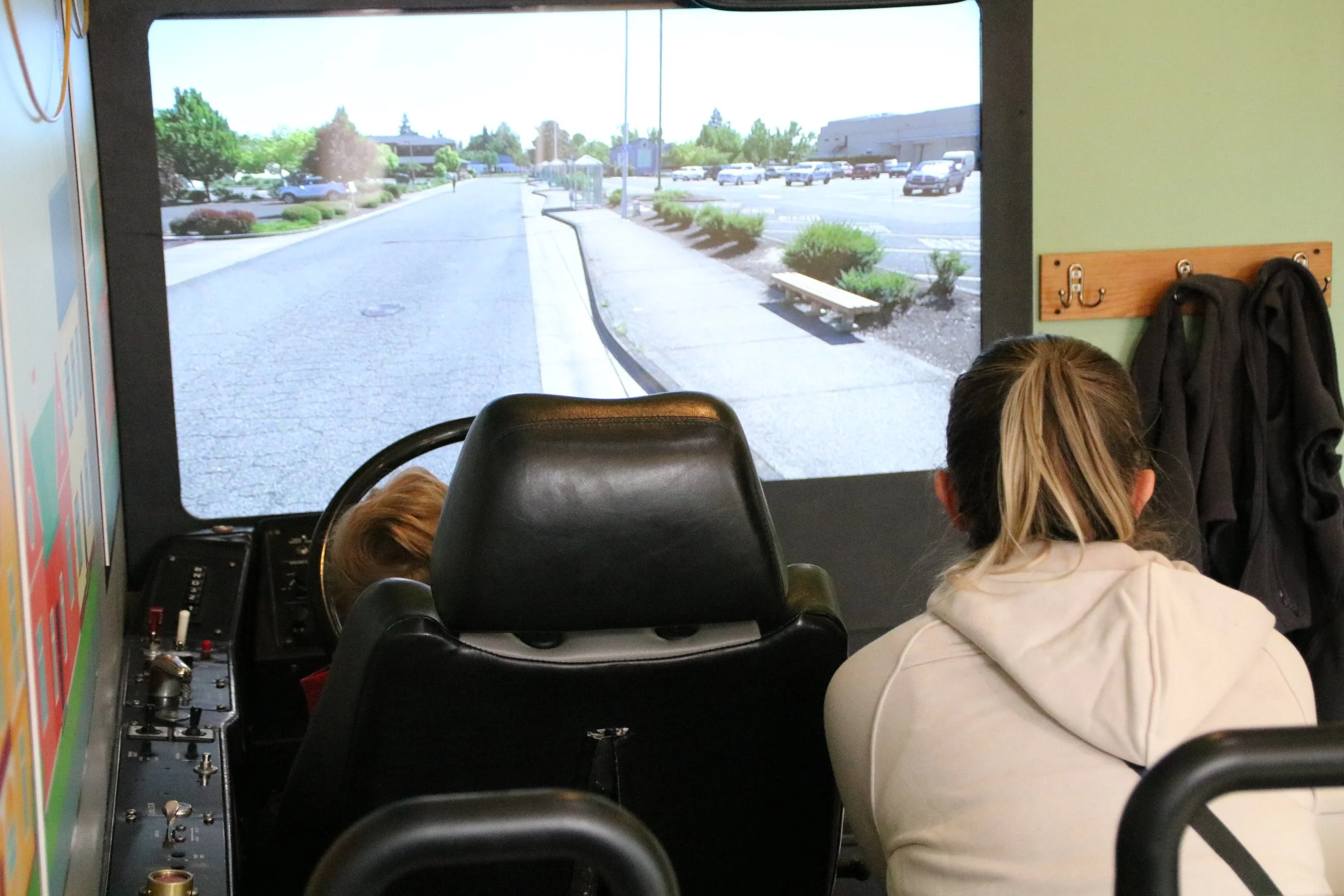 Children observe a parking lot and sidewalk on a large screen, with one child wearing a white hoodie in the foreground and a control panel on the left side.