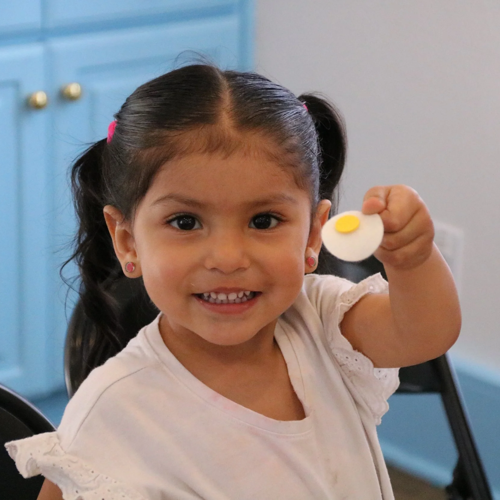 Young girl with dark hair in pigtails holding up a toy fried egg, smiling at the camera.