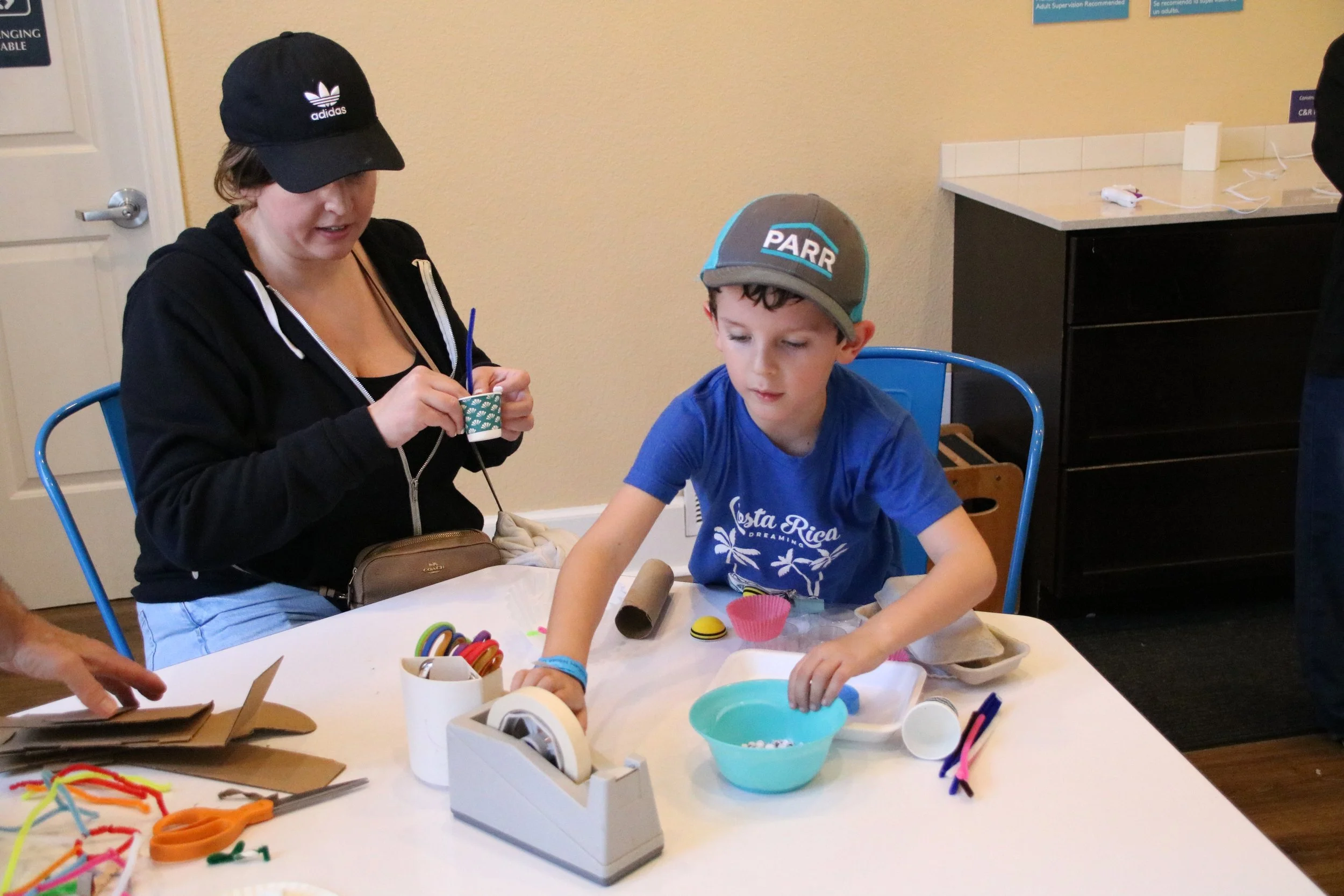 A boy wearing a blue Costa Rica t-shirt and a grey cap, sitting at a table, reaching into a bowl of small items. A woman nearby is weaving with pipe cleaners, with various craft supplies on the table.