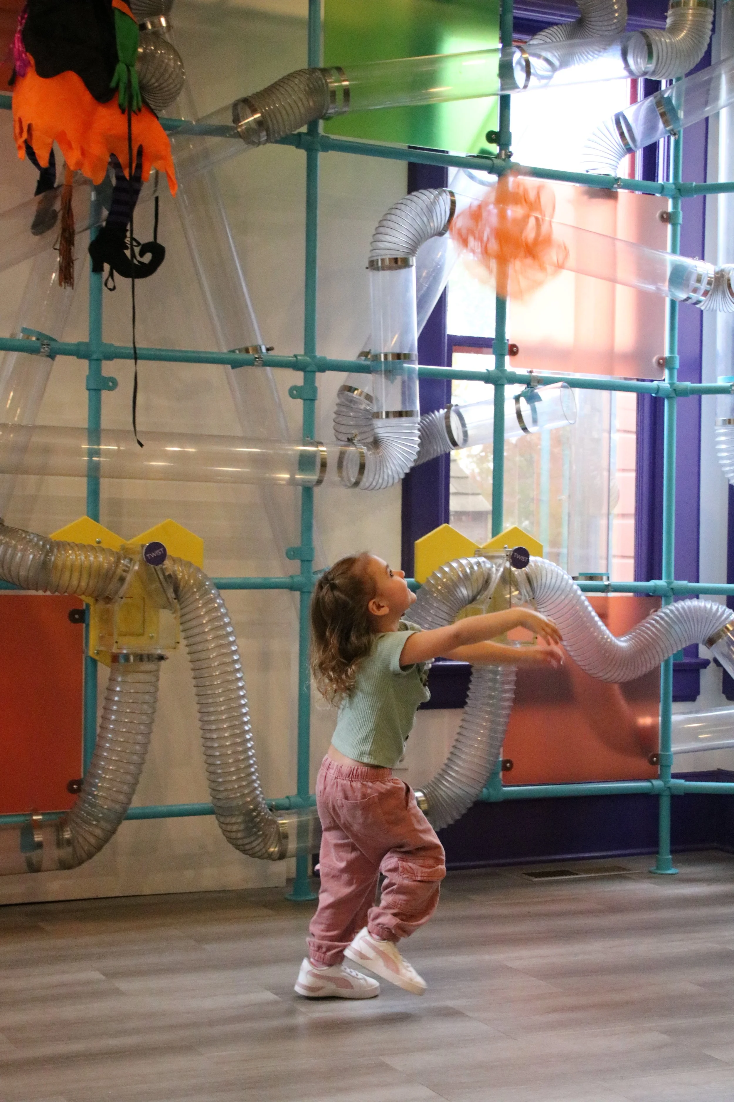 A young girl with curly hair, wearing a light green shirt, pink pants, and white sneakers, playing inside a colorful indoor playground with transparent tubes and orange decorations.