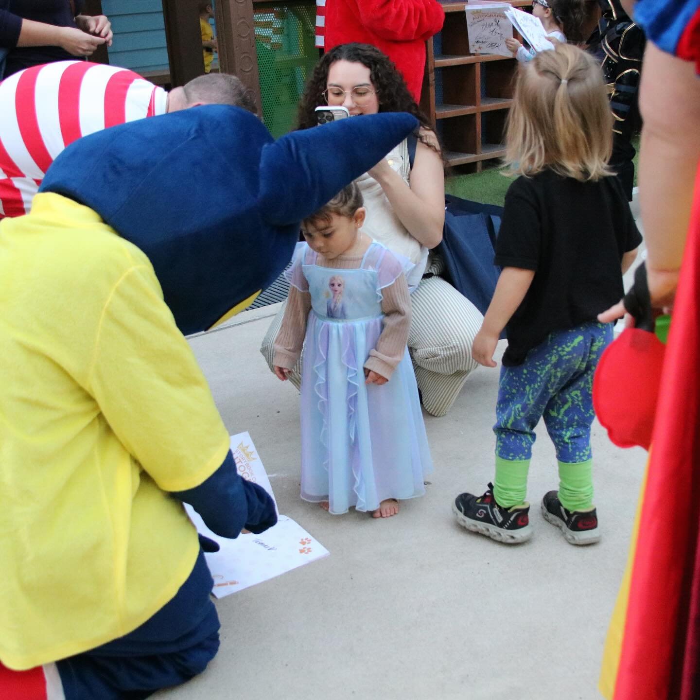 Pete the Cat mascot helps a young girl in an Elsa dress with a museum scavenger hunt map outdoors.
