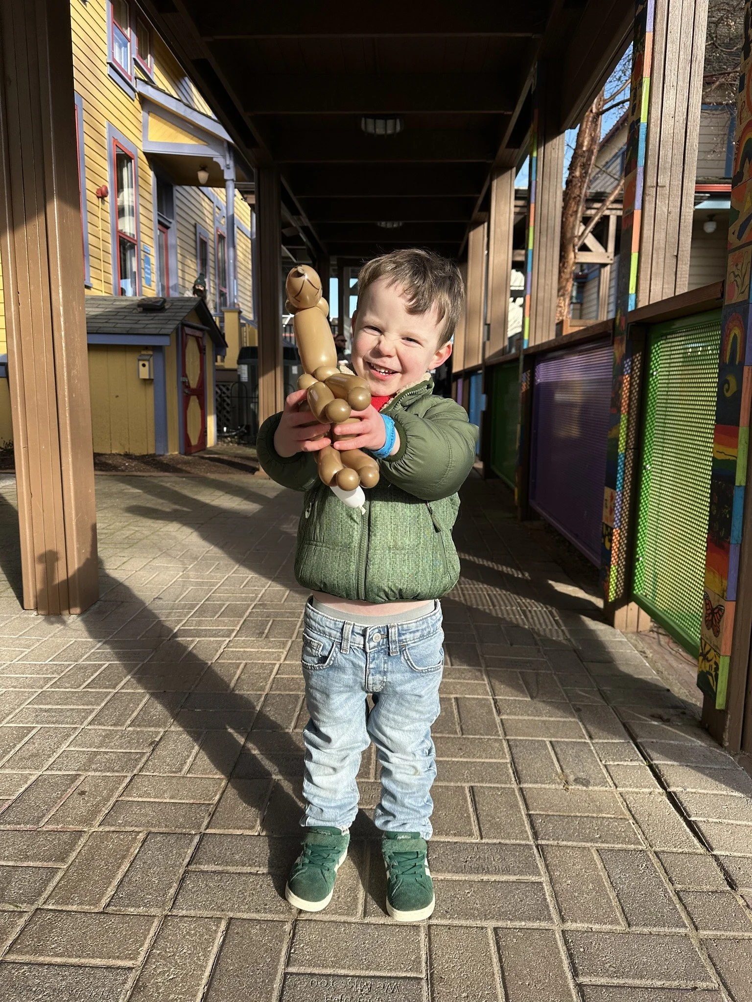 A young boy smiling and squinting in sunlight, holding a brown balloon animal puzzle. He is wearing a green jacket, light blue jeans, and green shoes, standing on a brick-paved walkway outside a colorful building.