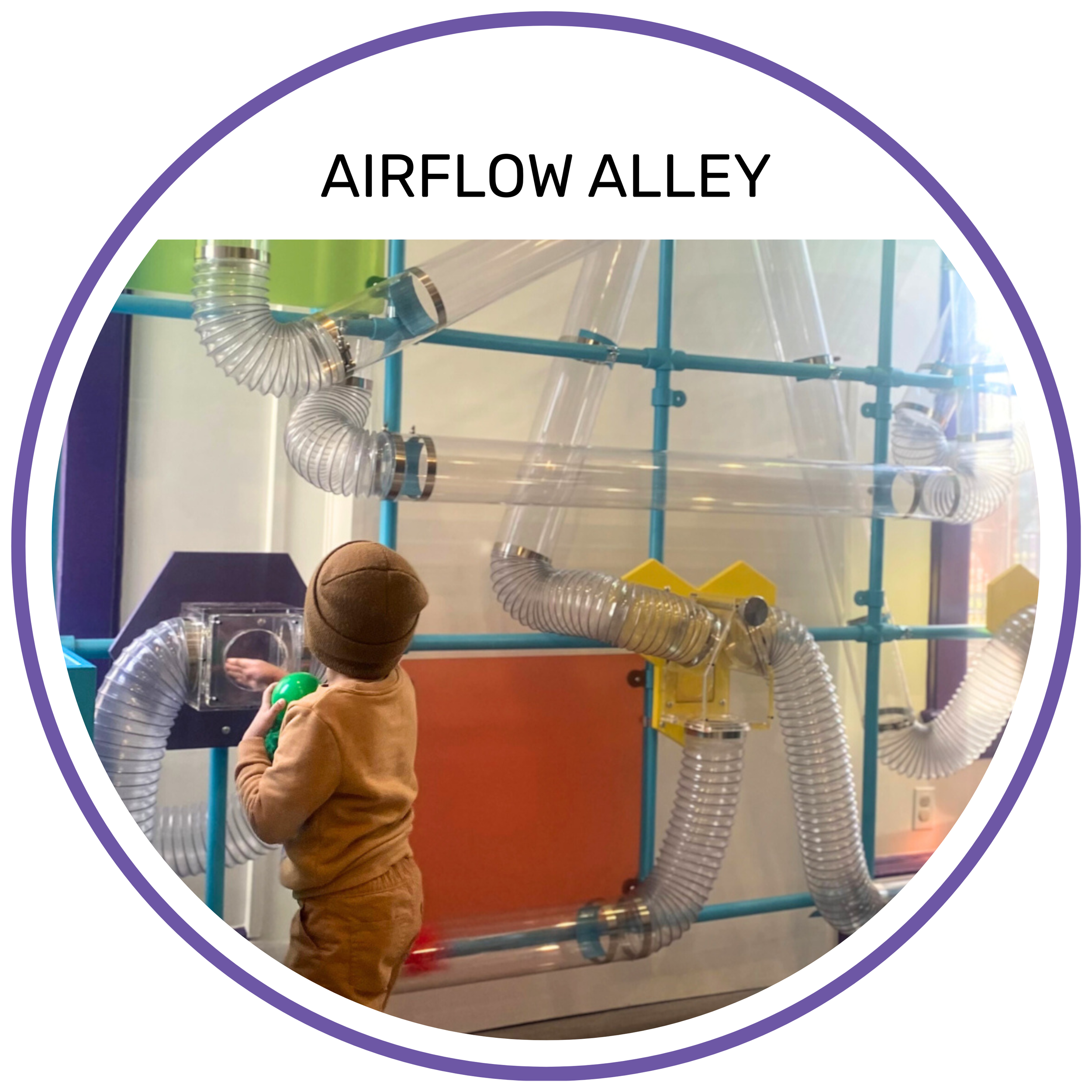A child in a brown outfit and hat interacting with a transparent interactive exhibit labeled 'Airflow Alley' that features a complex network of clear plastic tubes and blue support structures.