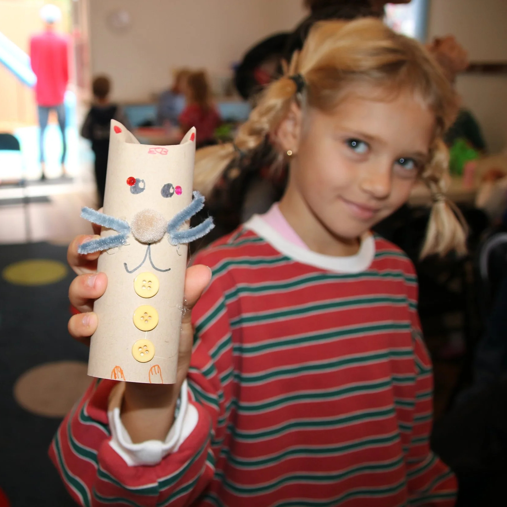A young girl with pigtails and a striped red and green shirt holds up a handmade craft resembling a mouse made from a paper toilet paper roll, decorated with buttons, pipe cleaner whiskers, and embellishments, in a classroom setting.