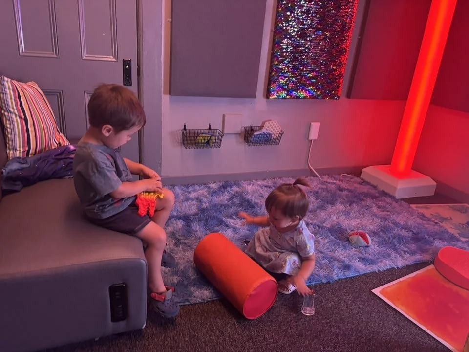 Two young children playing on a plush rug in a room with colorful lighting, toys, and wall decorations.