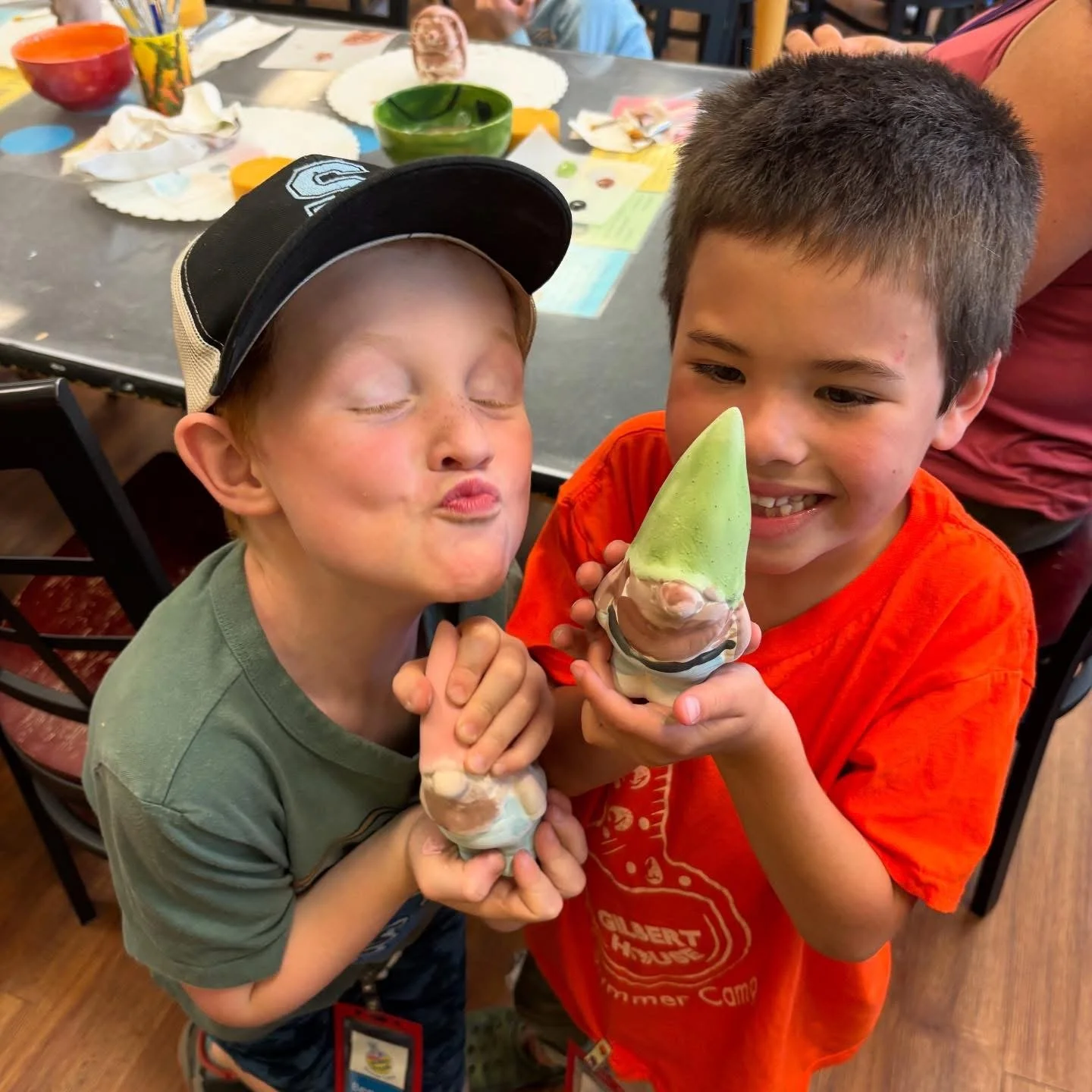 Two young boys holding ice cream cones and smiling. One boy is making a puckered face and wearing a baseball cap, while the other is smiling and wearing an orange T-shirt. They are at a table with bowls and papers in the background.