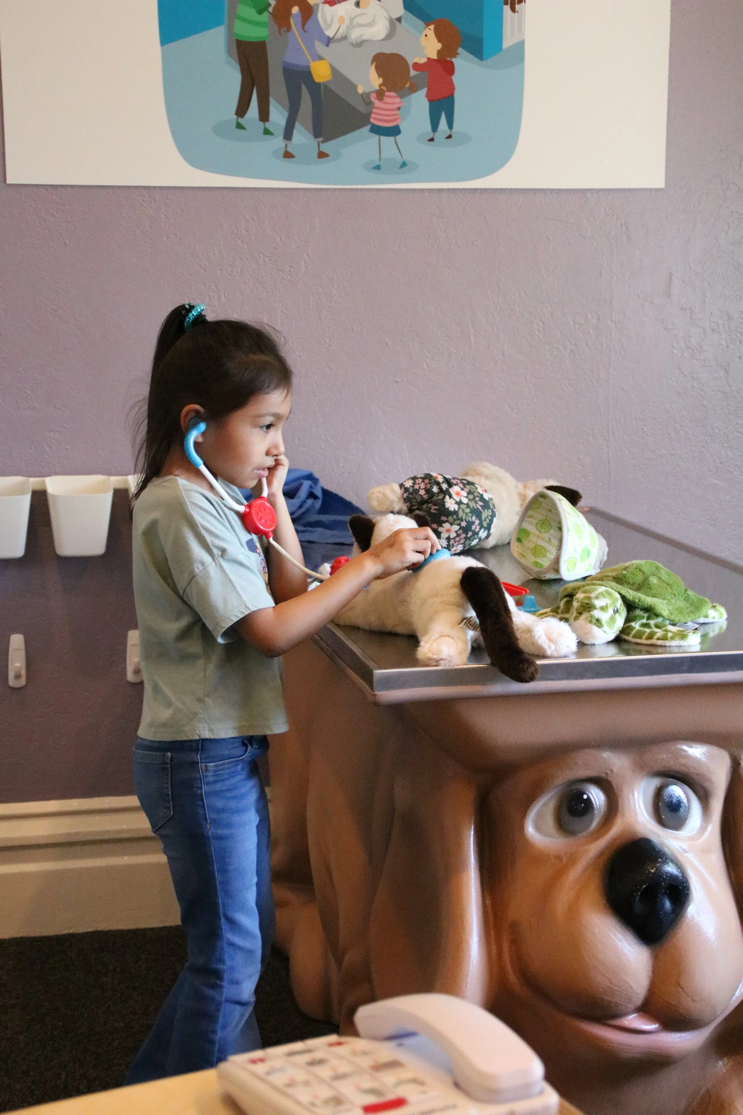 A young girl with a stethoscope checking a stuffed dog toy at a veterinary clinic, with other stuffed animals and toys on the table, and a large dog face sculpture at the counter.