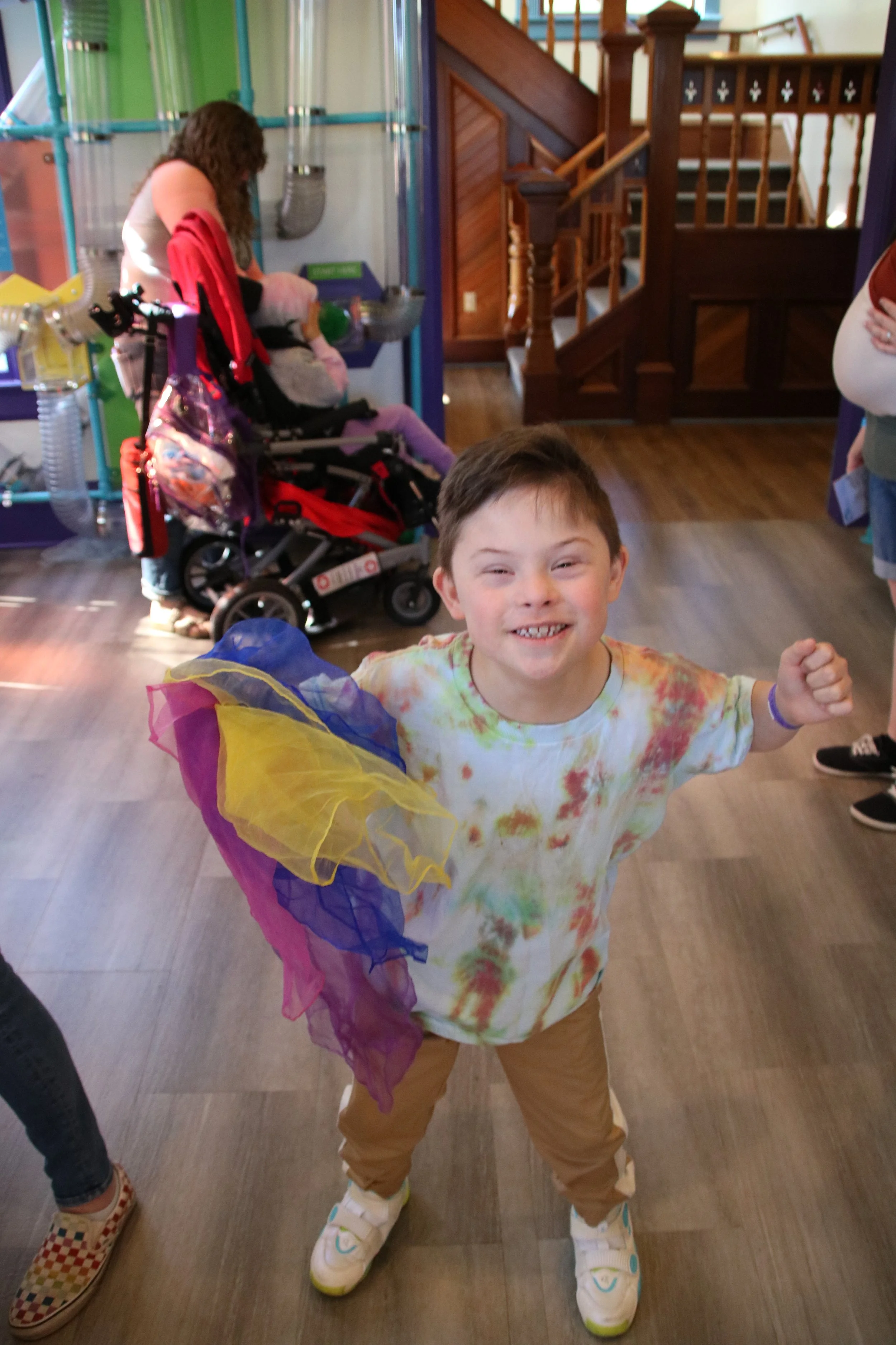 A young boy in a tie-dye shirt holding colorful fabric pieces, smiling excitedly at an indoor play area with wooden stairs and a woman in the background with a stroller.