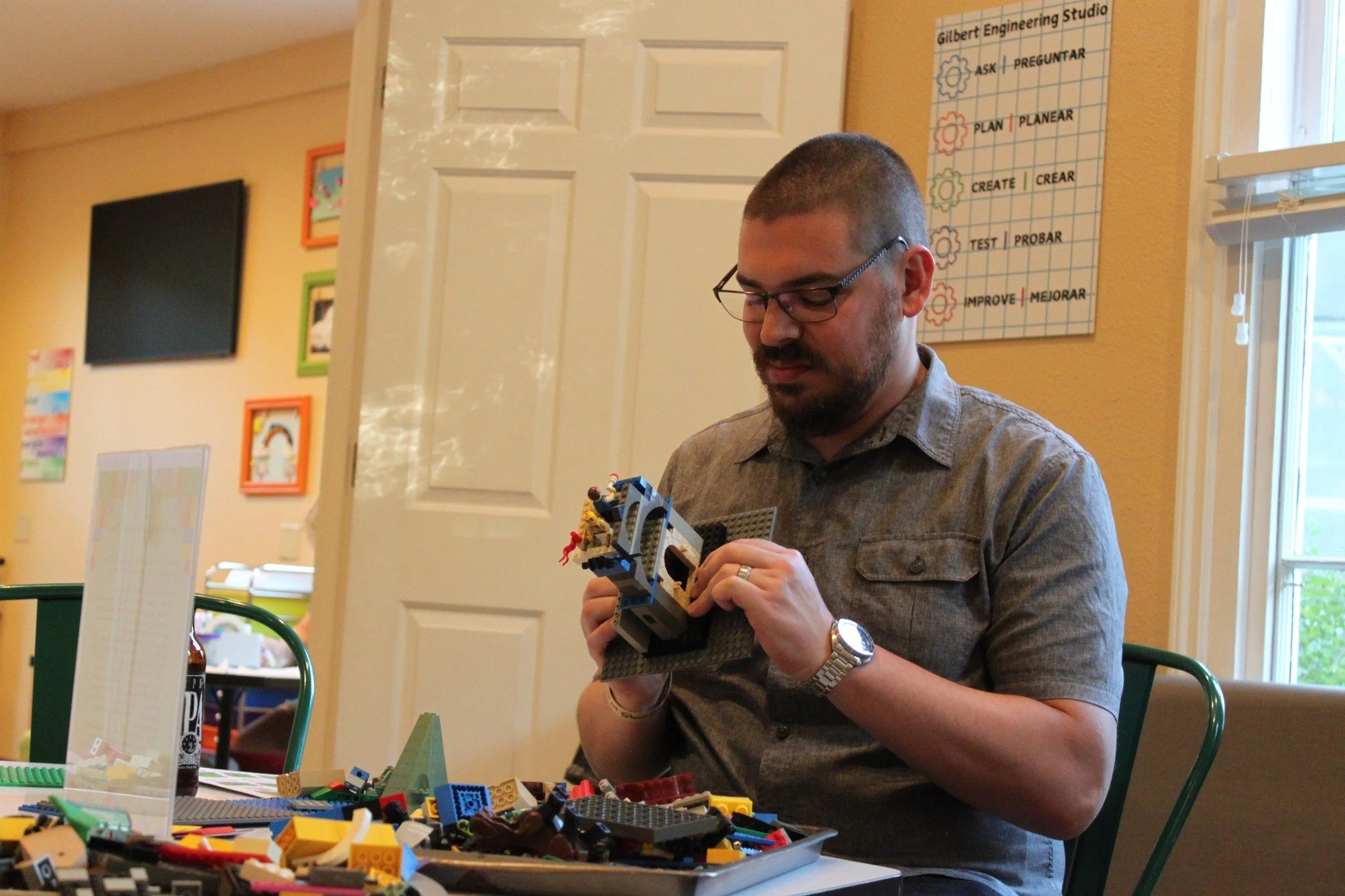 A man with glasses and a wristwatch, sitting at a table, assembling a LEGO set. The table is scattered with various LEGO pieces, and the man is focused on his building project in a room with colorful wall decorations and a poster on the wall.