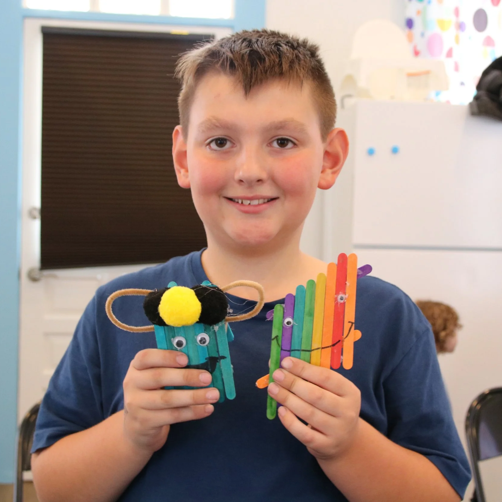 A smiling young boy holding two colorful craft projects made of rainbow-colored popsicle sticks, eyes, and craft materials, in a classroom setting.