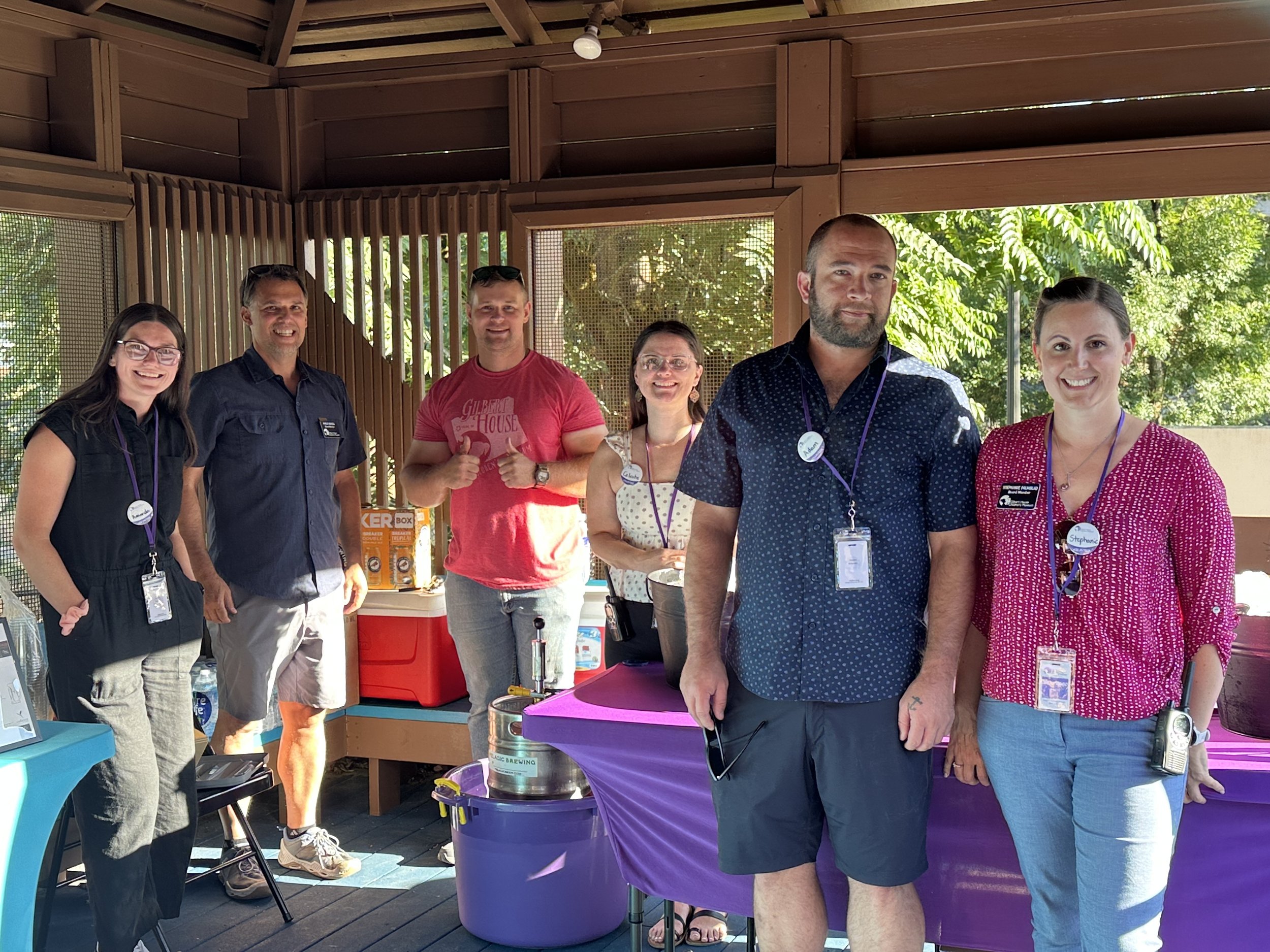 Group of six people standing inside a wooden shelter with trees outside. They are smiling, wearing ID badges, and standing near a table with a purple tablecloth and a drink dispenser.