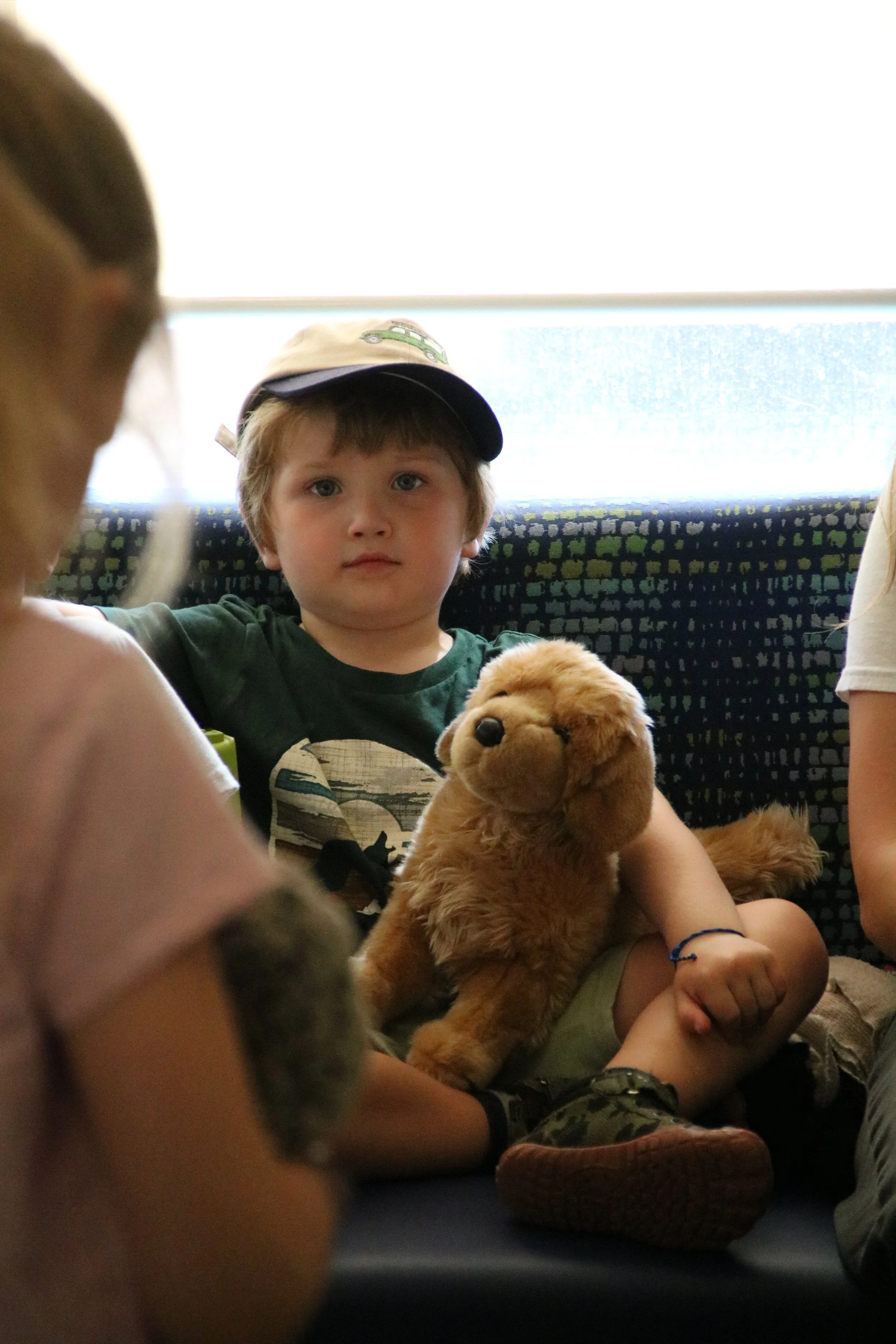 A young boy with a cap sitting on a bus holding a stuffed dog toy, with other children around him.