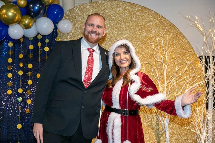 A man in a black suit and red tie standing next to a woman dressed as Mrs. Claus in a red velvet outfit with white fur trim, against a festive backdrop with gold and blue balloons and a gold circle, celebrating the holidays.