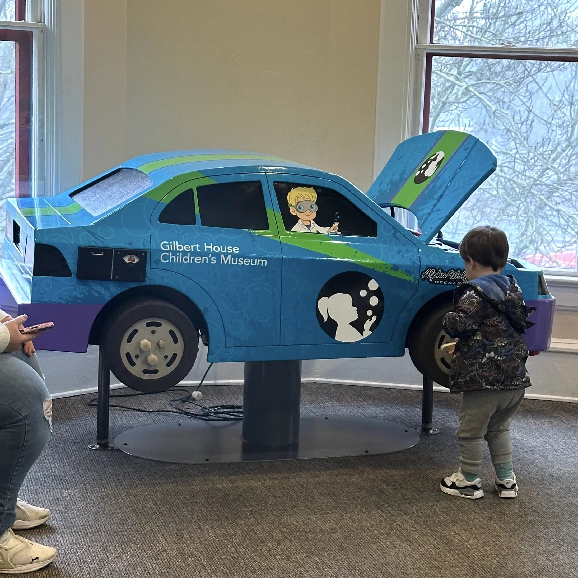 Children playing with a car-shaped interactive exhibit at Gilbert House Children's Museum, featuring a cartoon character in the driver's seat and a blue and green design.