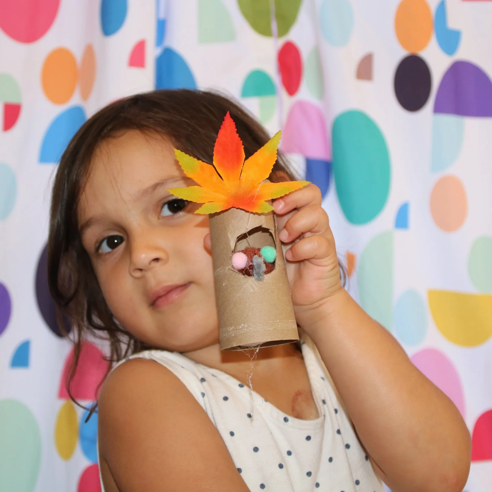 Child holding a cardboard tube decorated with a colorful autumn leaf and pom-poms, standing in front of a polka dot background.