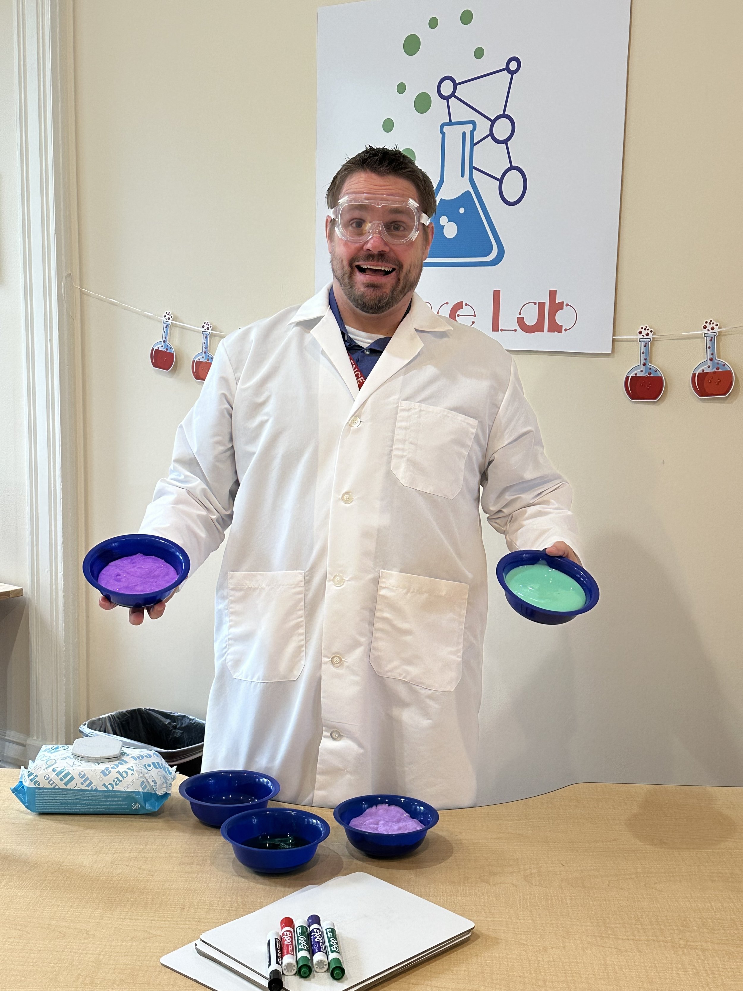 A man wearing a white lab coat and safety goggles is standing behind a table with bowls of colorful slime, holding two bowls of slime, one purple and one green, with a background featuring a science-themed banner and a string of paper cutouts of beakers.