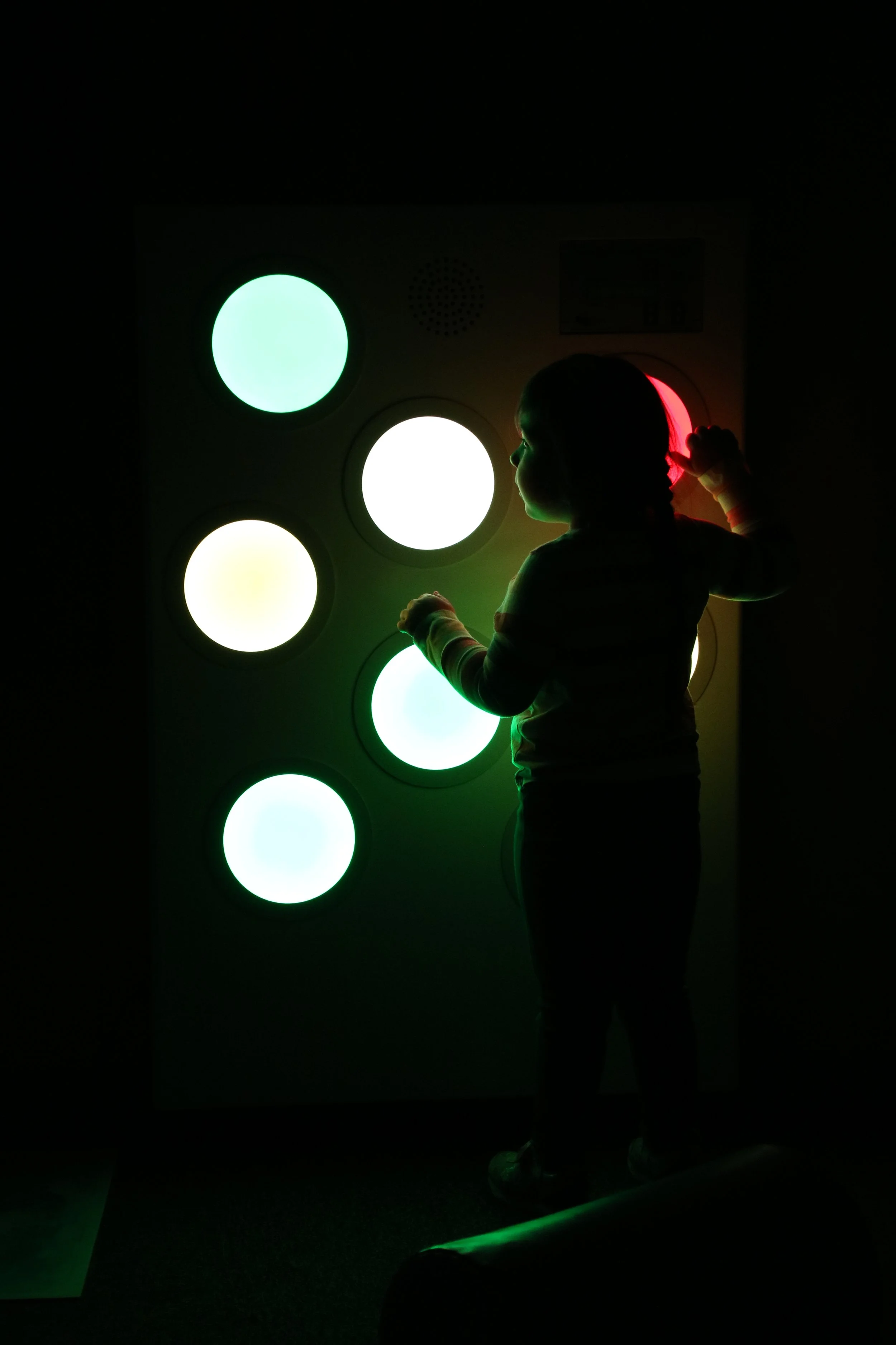 Child standing in front of an interactive light display with glowing circles of different colors, including green, white, yellow, and red.