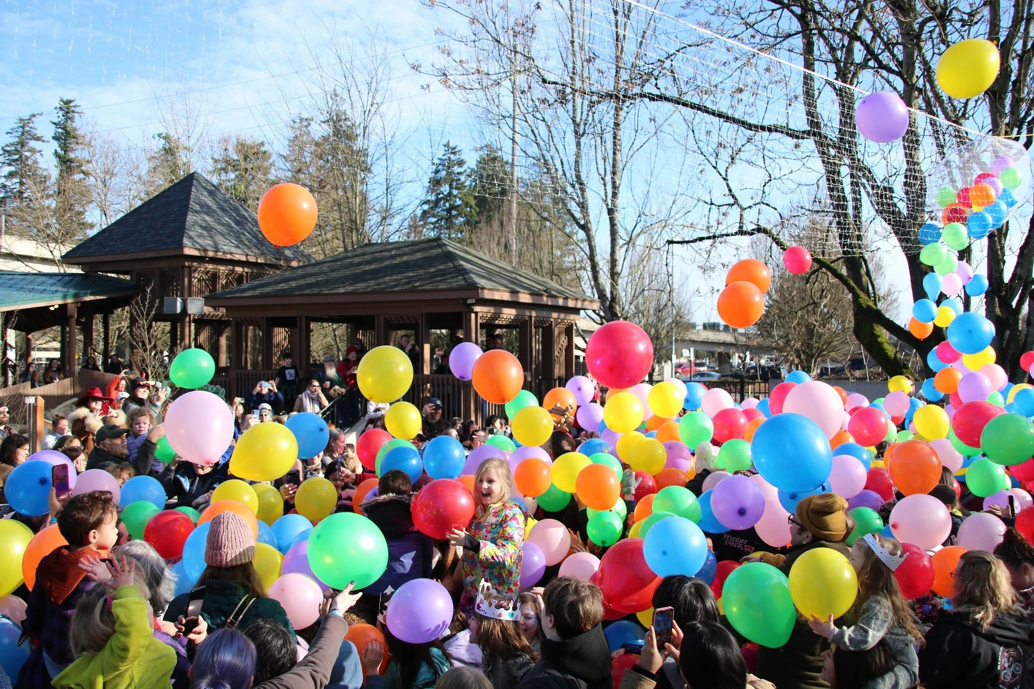 Children and adults participating in a balloon release event outdoors, with numerous colorful balloons in the air and a wooden pavilion in the background.