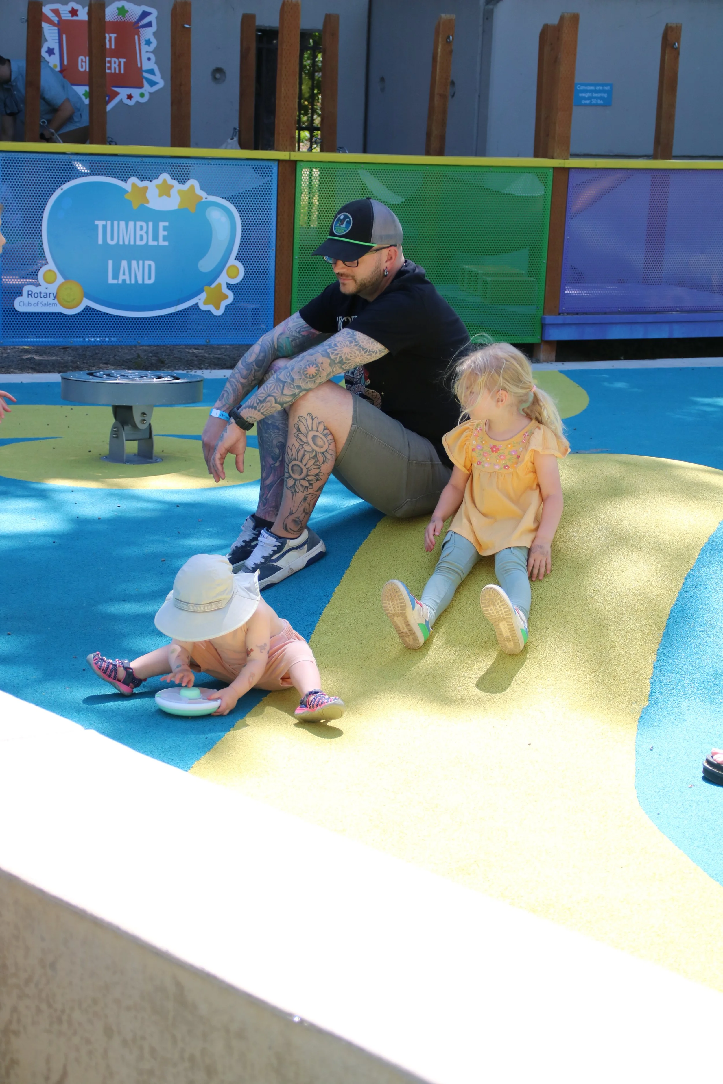 A man sitting on a colorful playground surface with two small children playing nearby. The playground sign reads "Tumble Land" and is decorated with stars. One child is wearing a large sunhat and pink outfit, reaching for a toy, while the other child is sitting against the man, wearing a yellow dress and gray leggings.