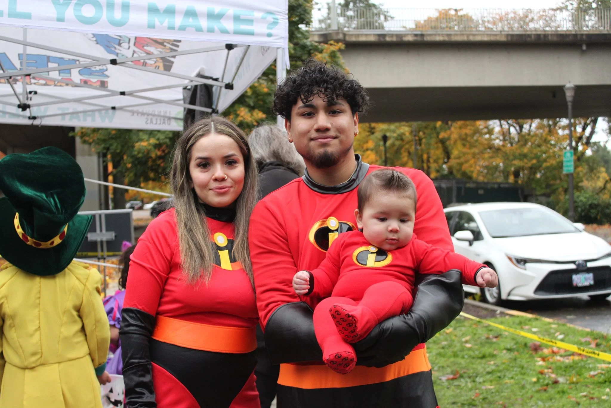 Family dressed as the Incredibles at a Halloween event, standing outdoors near a tent and parked cars with fall foliage in the background.
