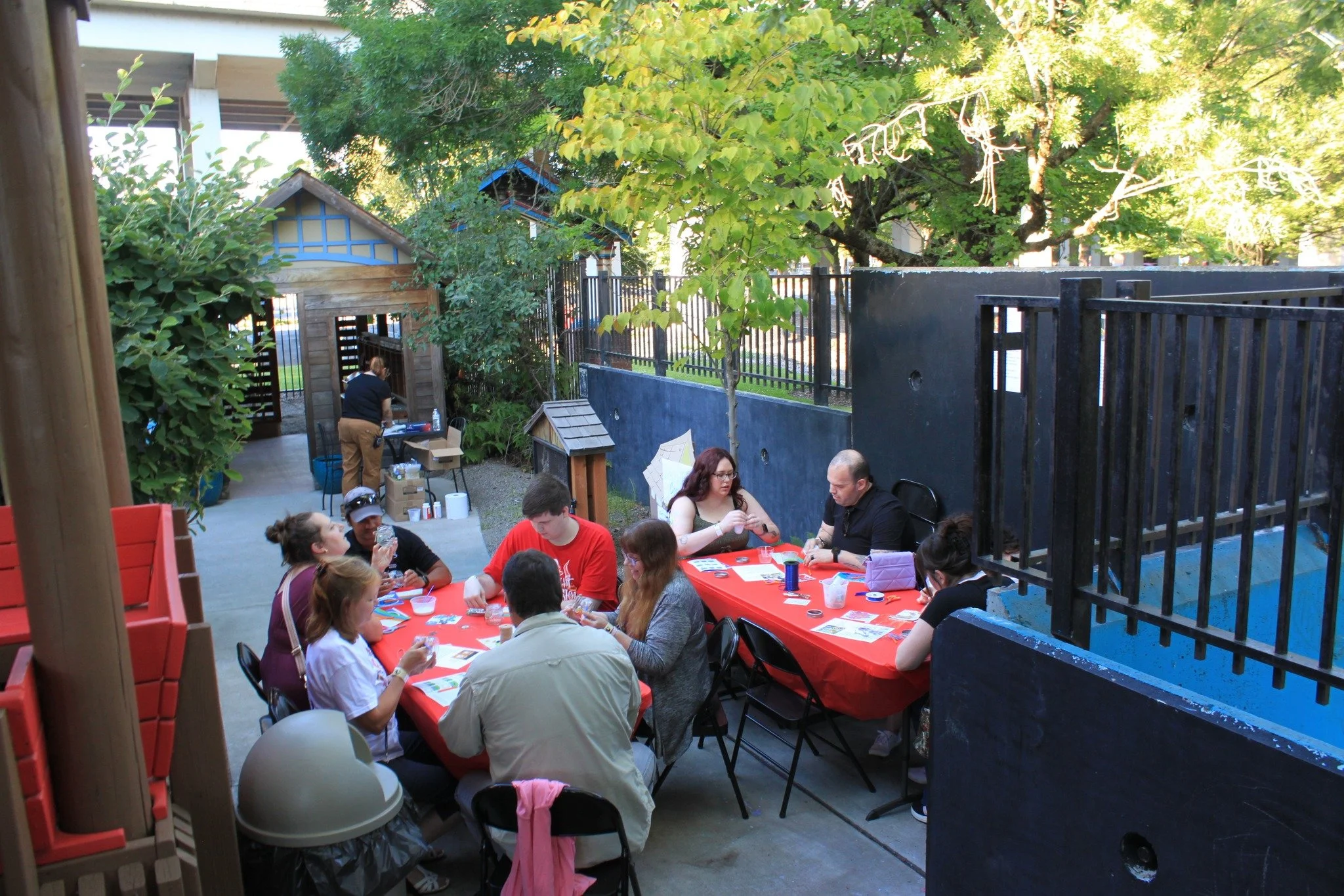 A group of people gathered around a red tablecloth covered table outdoors, engaging in activities like eating or playing games, with trees and a small wooden playhouse nearby.
