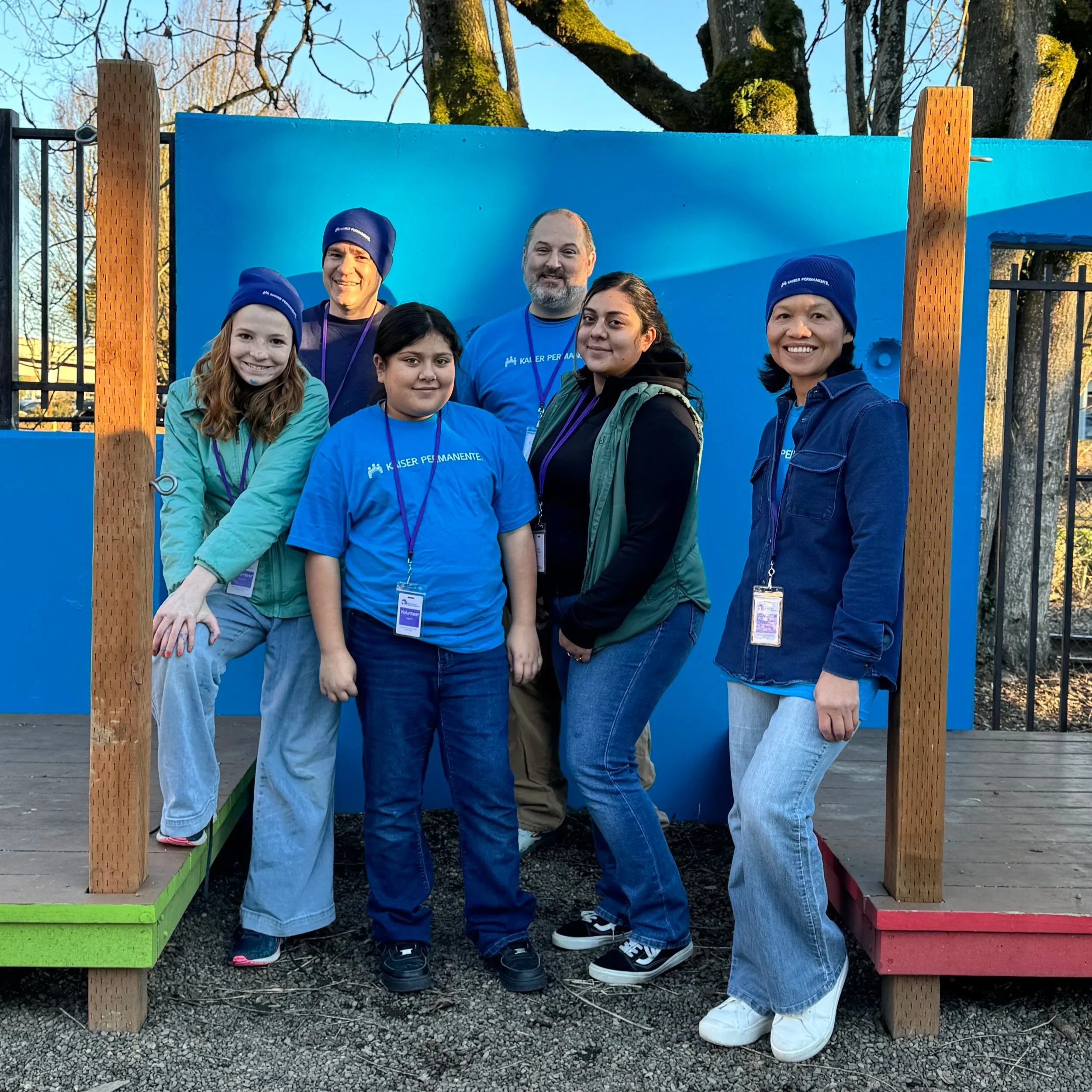 Group of six diverse people standing in front of a bright blue wall with wooden posts at an outdoor event during daytime.